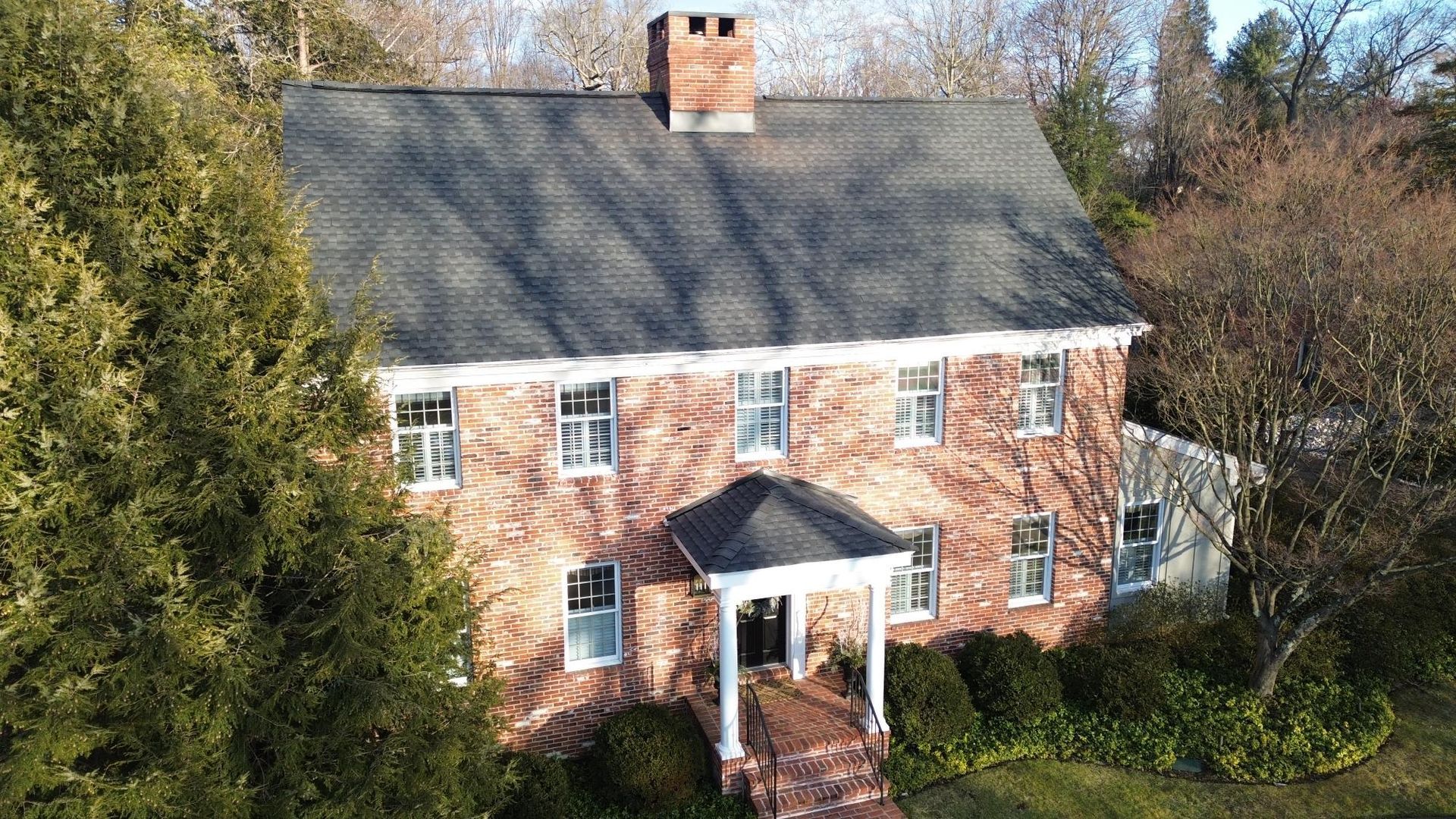 Two-story brick house with a dark roof and a front porch, surrounded by trees and greenery.