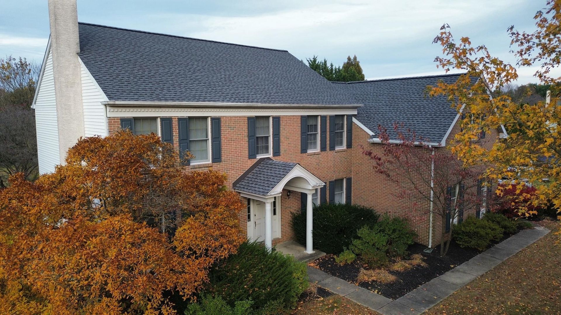 Brick house with dark roof and white chimney, surrounded by trees with fall foliage.