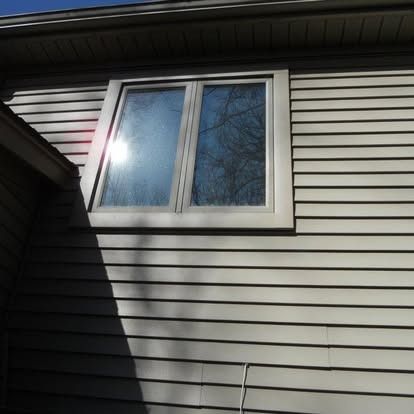 Exterior wall with a window, siding is horizontal and beige. Sunlight reflects off the window glass.