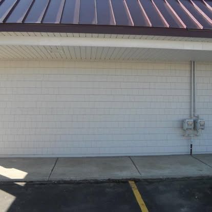 Exterior of a building with white siding, brown roof, and concrete sidewalk. Electrical conduit runs up the wall.