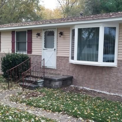 Ranch-style house with tan siding, stone facade, and bay window. Front door with storm door and small porch.