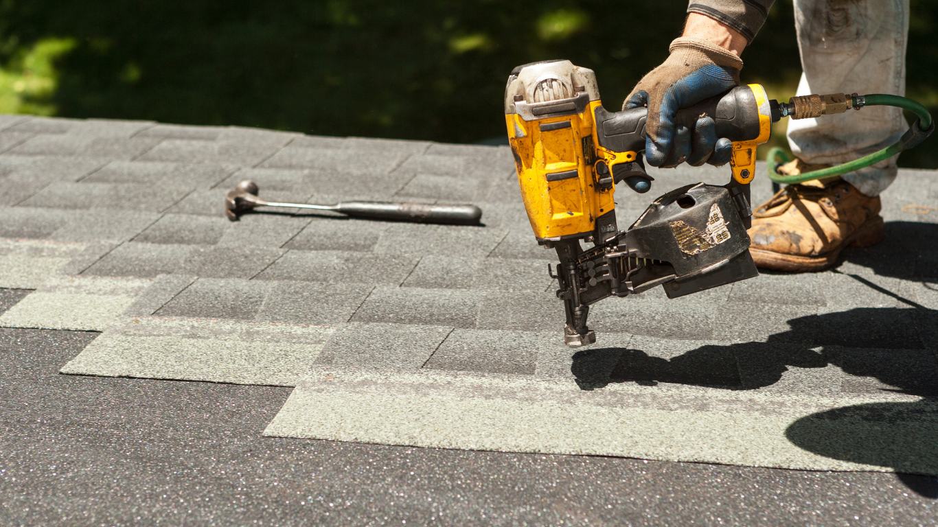 Roofer using a nail gun to install shingles on a roof. A hammer is nearby. Sunny outdoor setting.