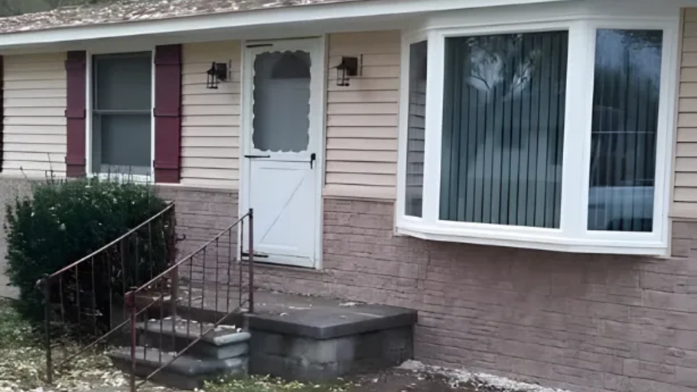 Ranch-style house with tan siding, stone facade, and bay window. Front door with storm door and small porch.