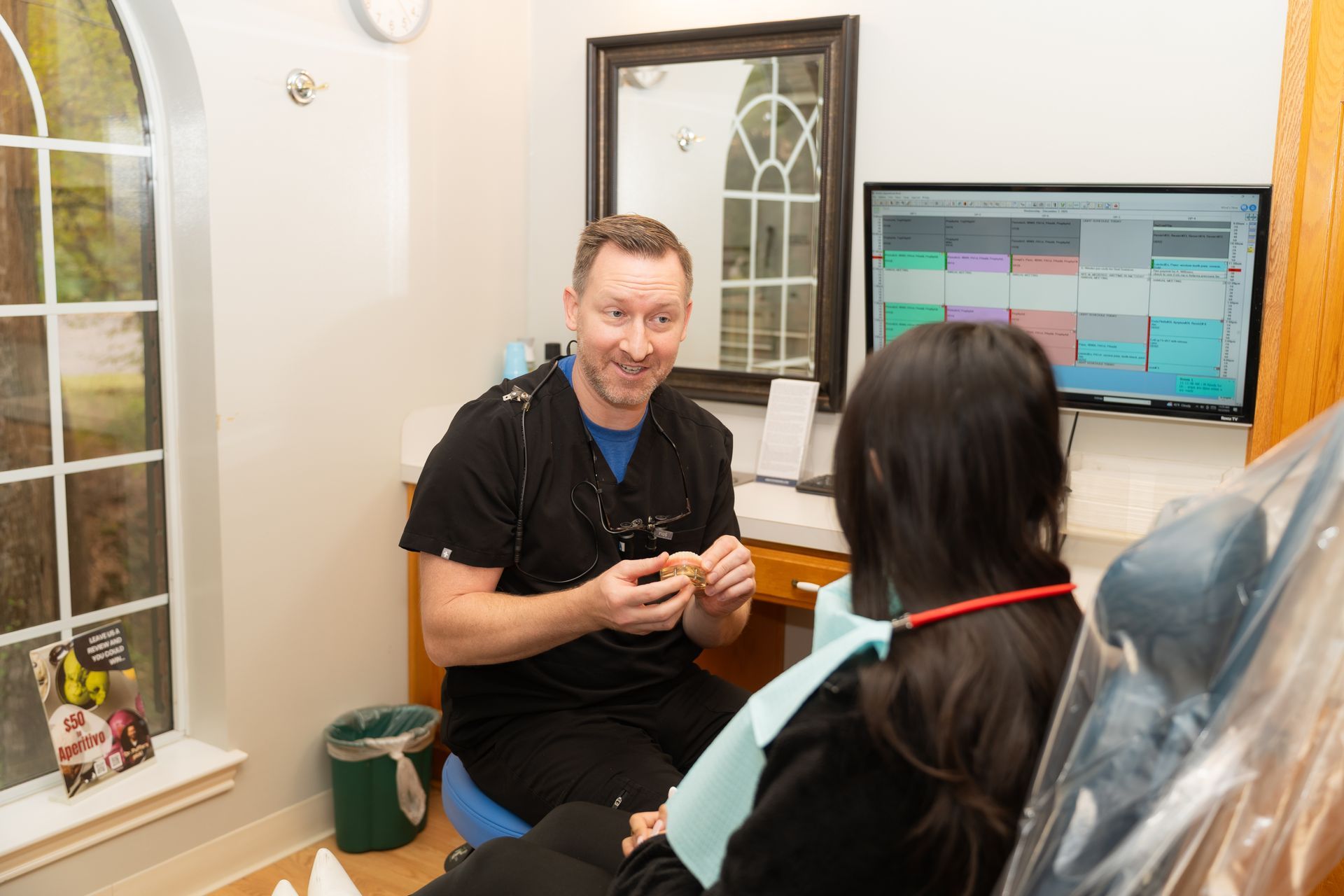 A dental professional holds a small model while speaking with a patient seated in an exam chair in an office setting.