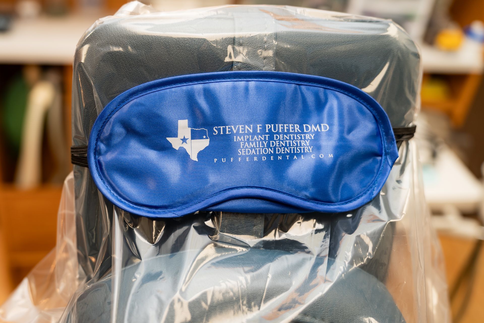 A blue sleep mask with a white Texas silhouette and dentist branding sits on a plastic-covered dental chair.