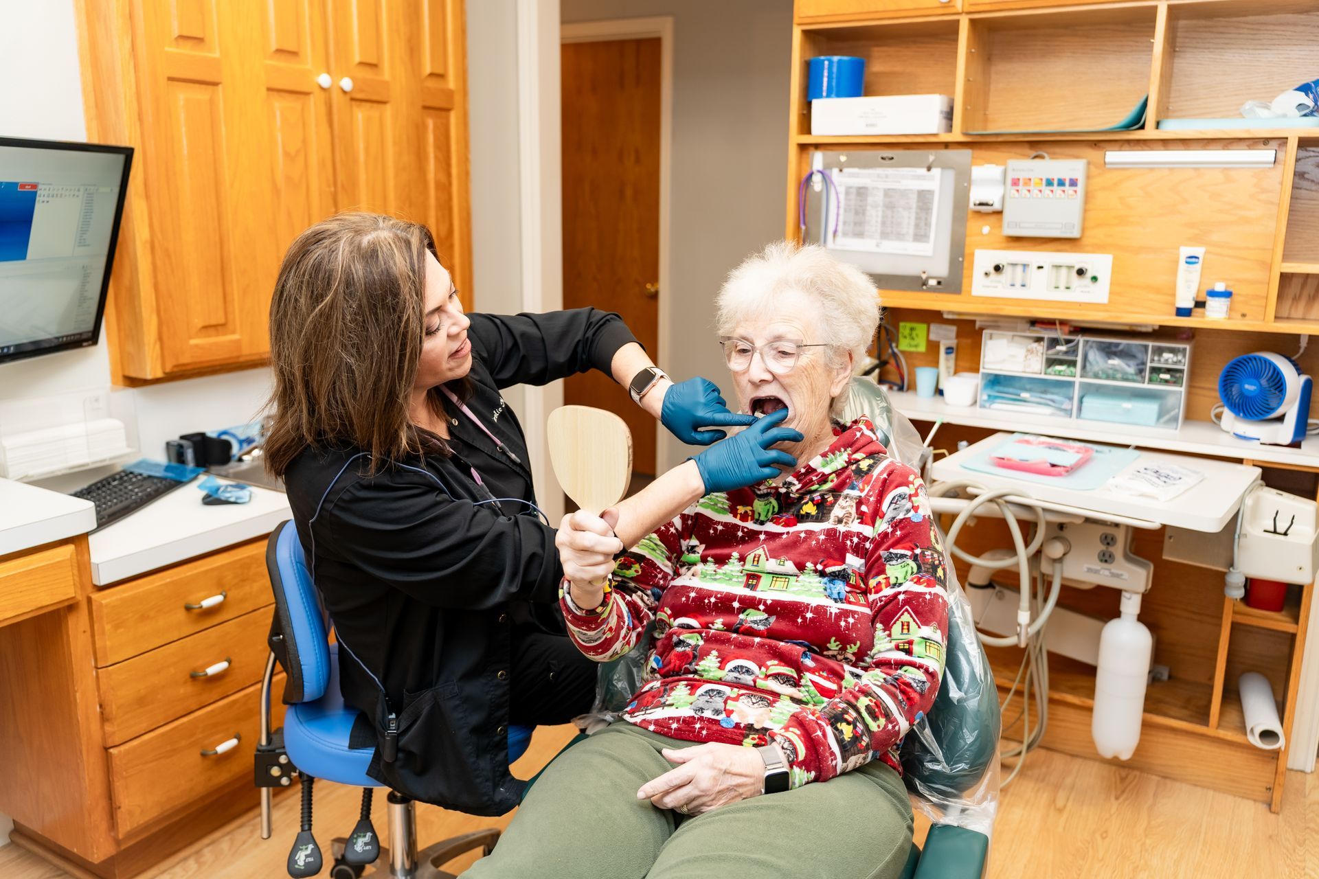 A dental professional in a clinic examines a patient's mouth while the patient holds a mirror to view the procedure.