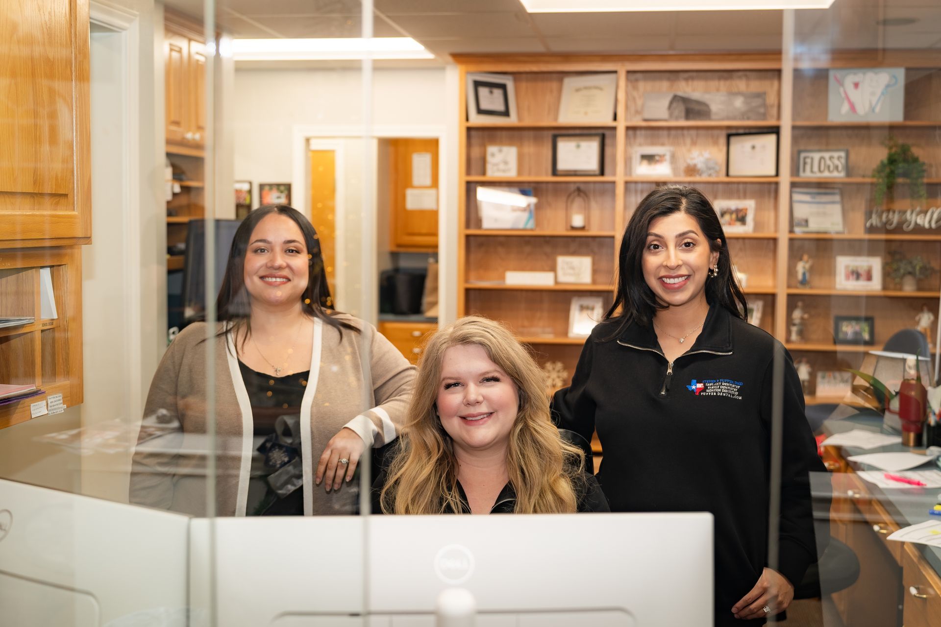 Three office staff members smiling and posing behind a front desk with computers in an office with wooden shelving.
