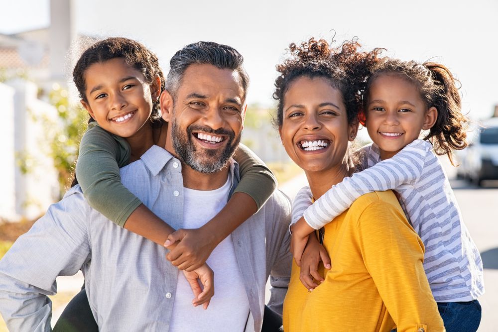 Two parents smiling while giving their two children piggyback rides outdoors on a sunny day.