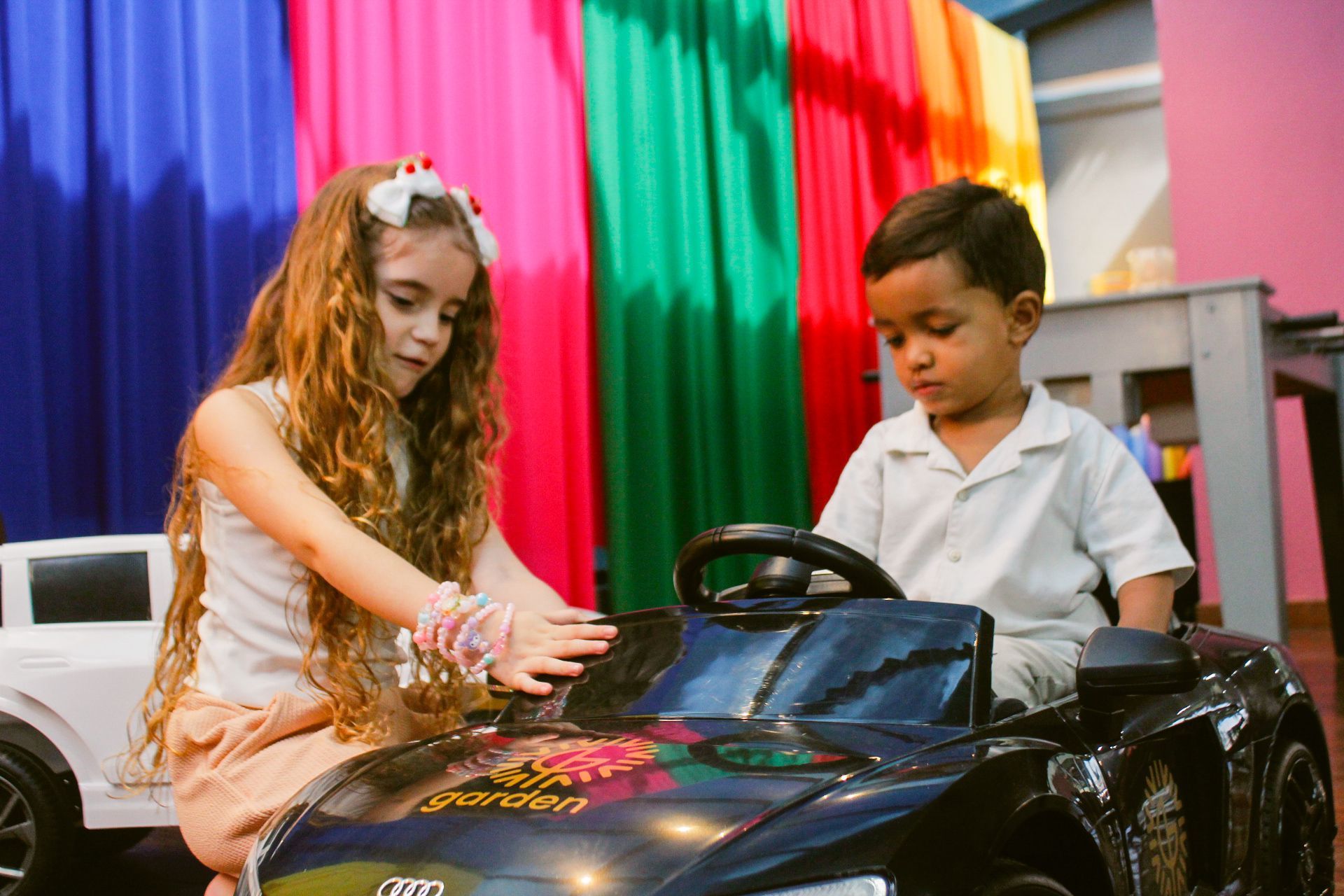 Menina e menino brincando com um carrinho de brinquedo em frente a um tecido colorido drapeado.