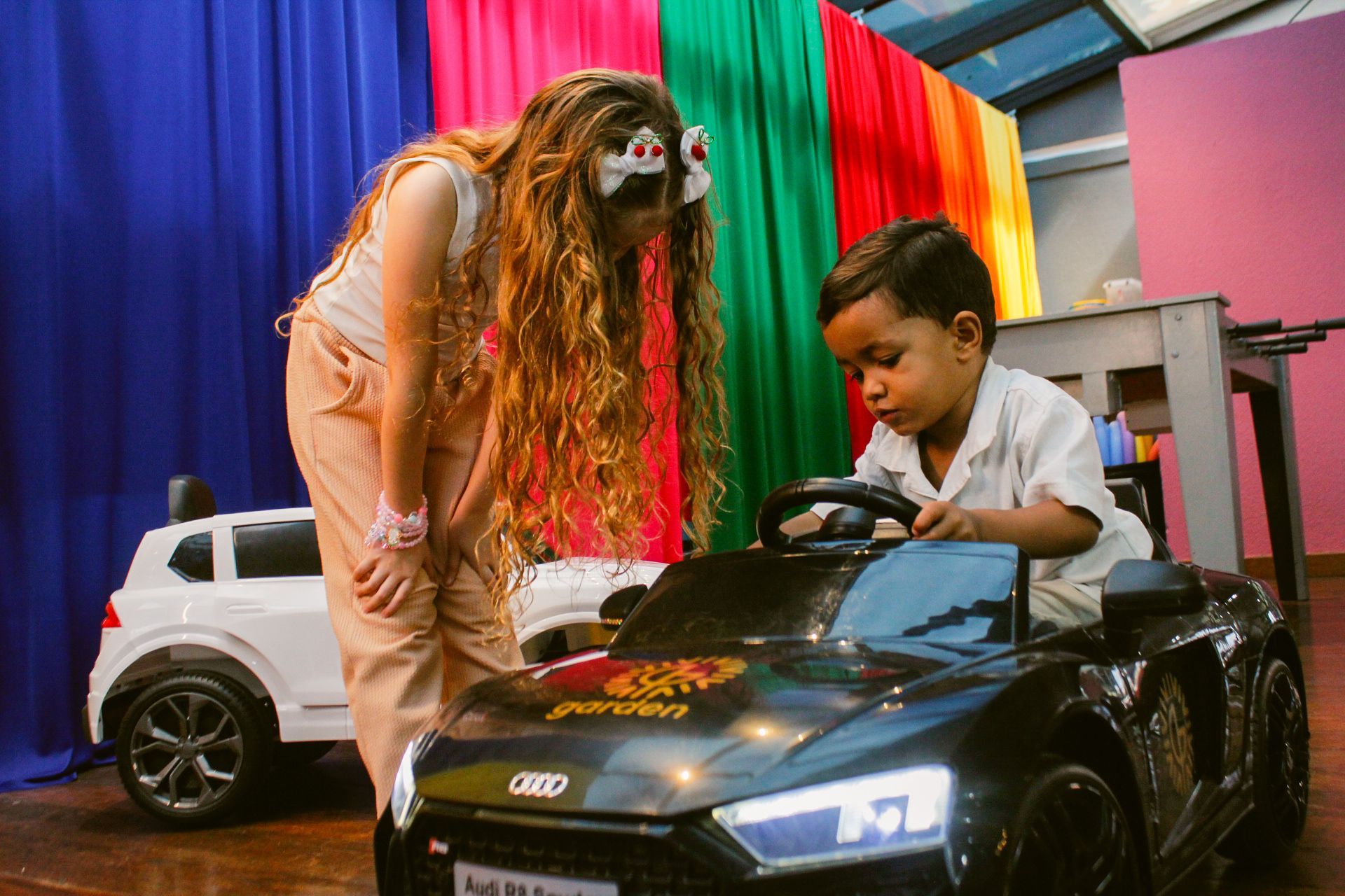 Mulher se inclina para observar uma criança em um carrinho de brinquedo. Cenário colorido.