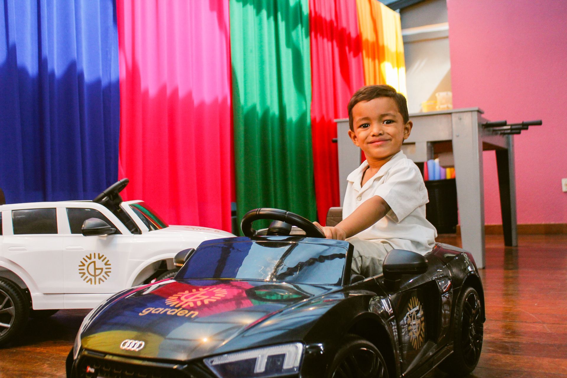 Menino de camisa branca sorri enquanto está sentado em um carrinho de brinquedo preto; um carrinho de brinquedo branco está à esquerda, em um cenário colorido.