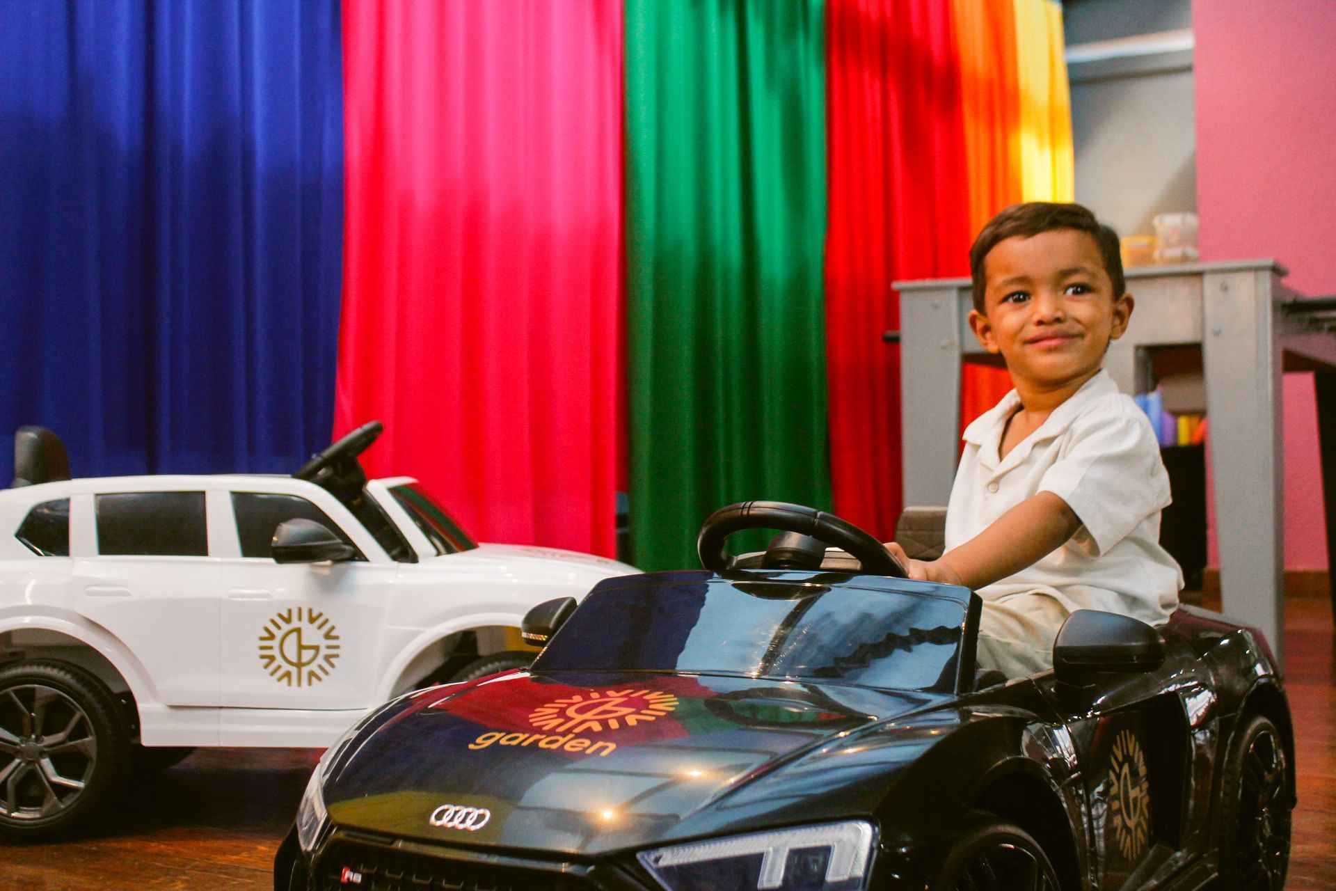 Menino sorrindo em um carrinho de brinquedo; outro carrinho de brinquedo atrás dele; cortinas coloridas ao fundo.