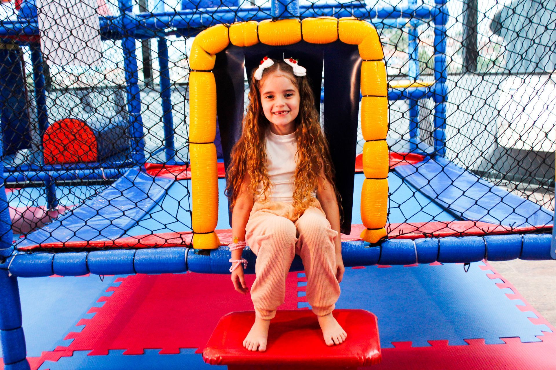 Menina sorri, sentada em uma estrutura colorida de um parque infantil coberto.