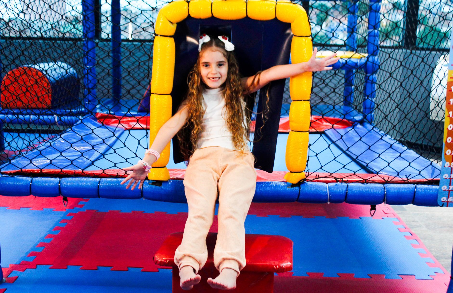 Menina sentada em um banco vermelho, com os braços estendidos, sorrindo em um parquinho infantil coberto.