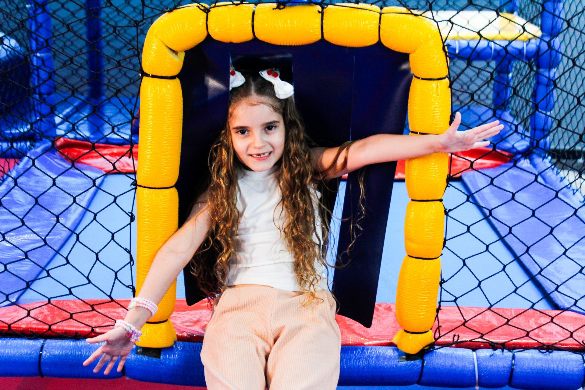 Menina com os braços estendidos, sorrindo de dentro de uma estrutura de brinquedo com moldura amarela, em ambiente interno.