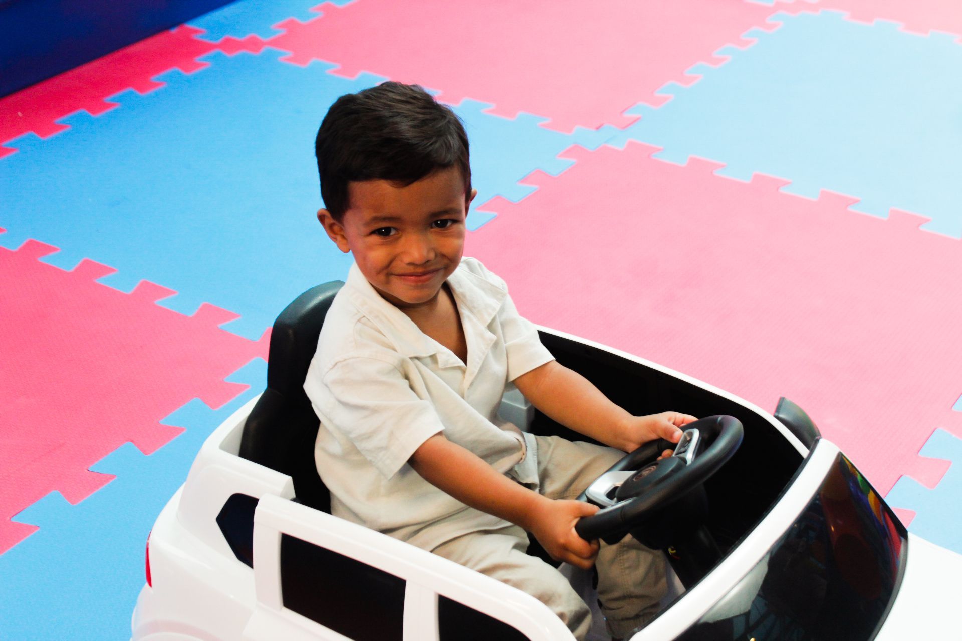 Criança sorrindo, dirigindo um carrinho de brinquedo branco em um piso rosa e azul.