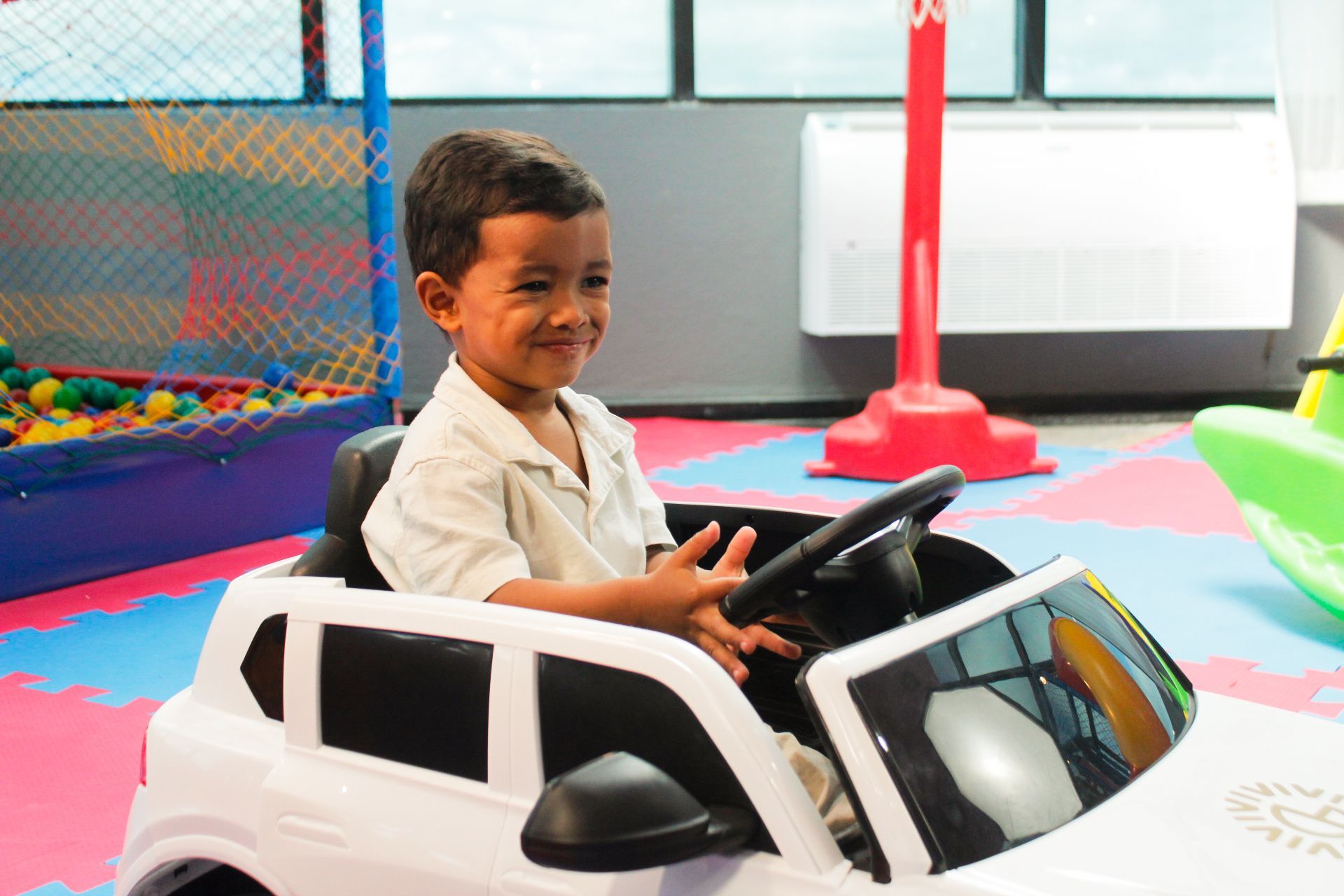 Menino sorri enquanto está sentado em um carrinho de brinquedo branco. Ele está em uma sala de jogos com piscina de bolinhas.