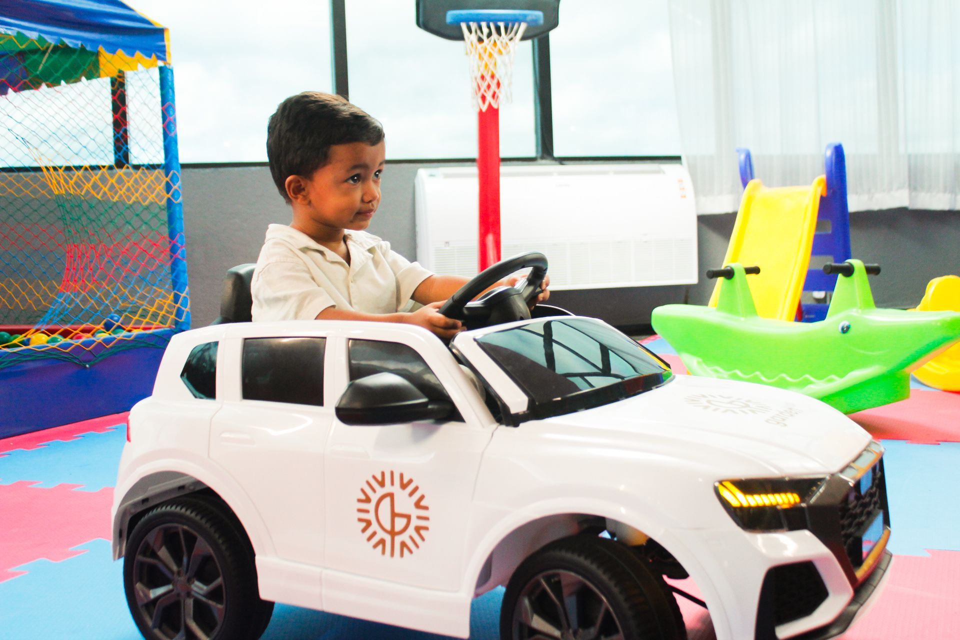 Criança pequena dirigindo um carrinho de brinquedo branco em uma sala de jogos, olhando para frente.