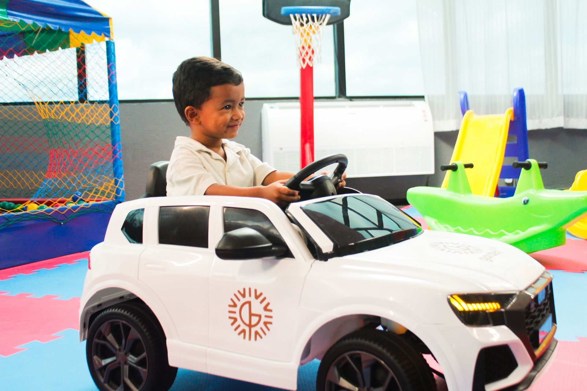 Criança pequena dirigindo um carrinho de brinquedo branco em uma sala de jogos com cesta de basquete e escorregadores.