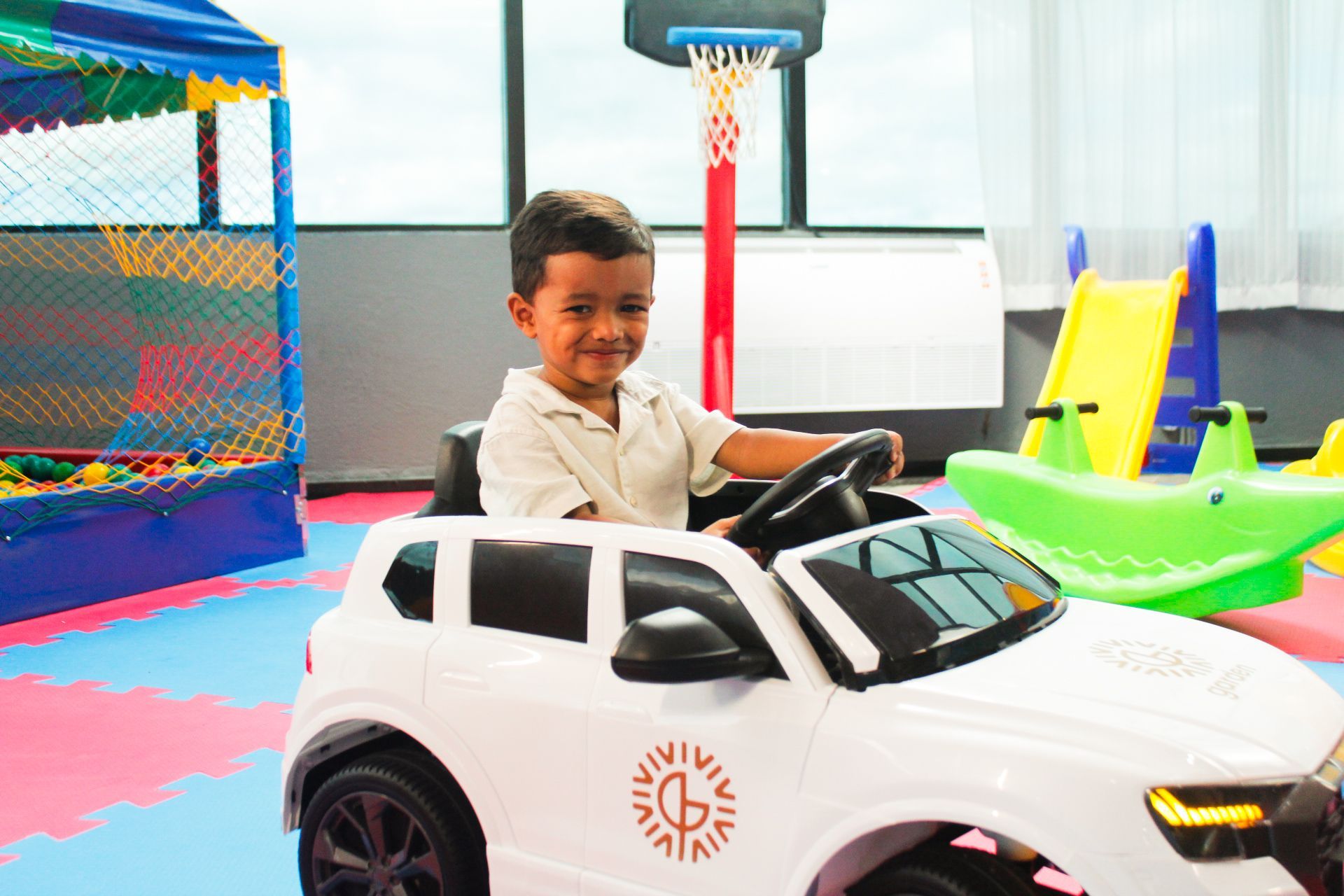 Menino sorrindo em um carrinho de brinquedo branco dentro de uma sala de jogos colorida.