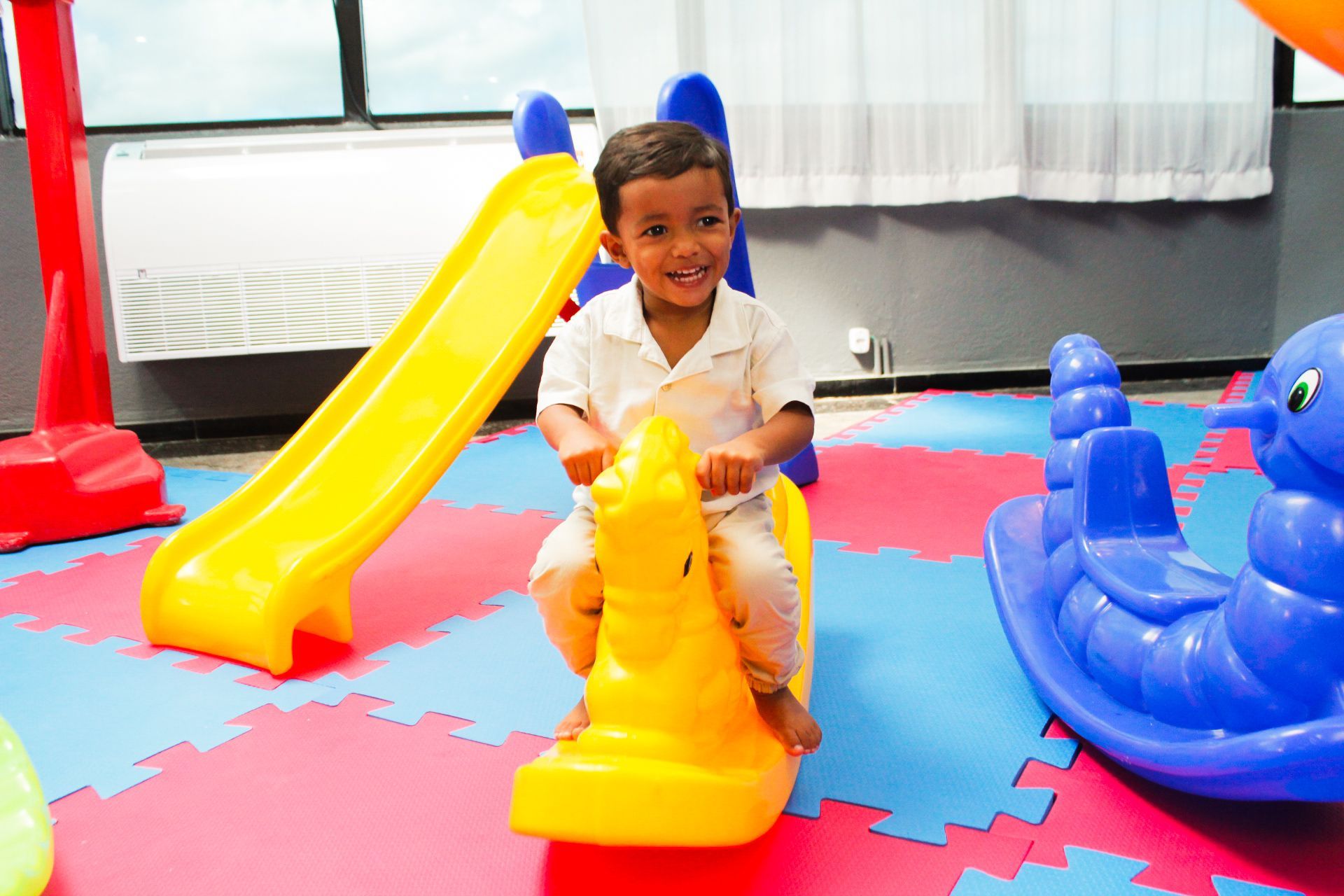 Menino sorrindo enquanto monta um cavalinho de plástico amarelo em uma área de recreação com escorregador e barco de balanço.