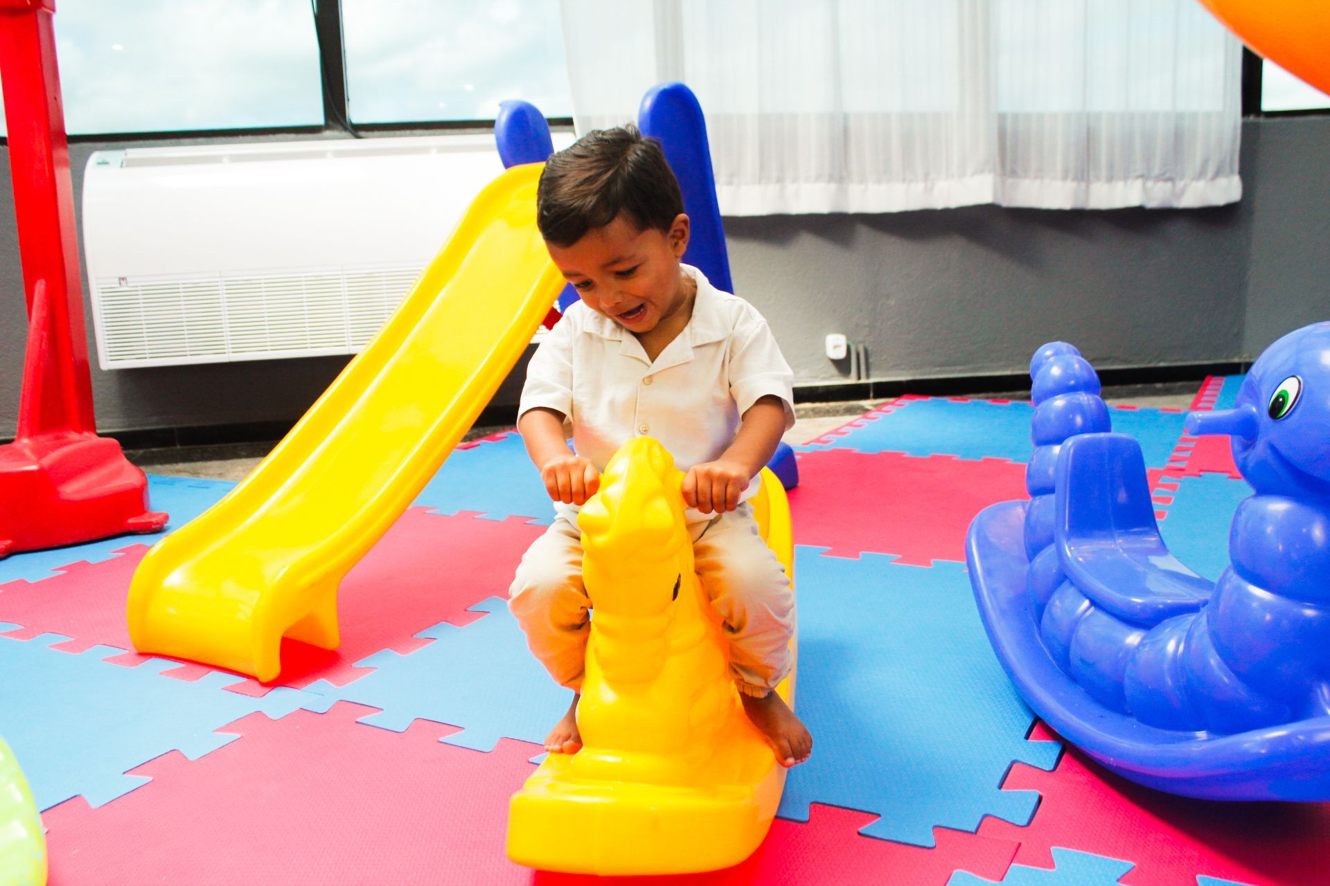 Criança montada em um cavalo de balanço amarelo em uma área de recreação com um escorregador amarelo e um brinquedo de balanço azul.