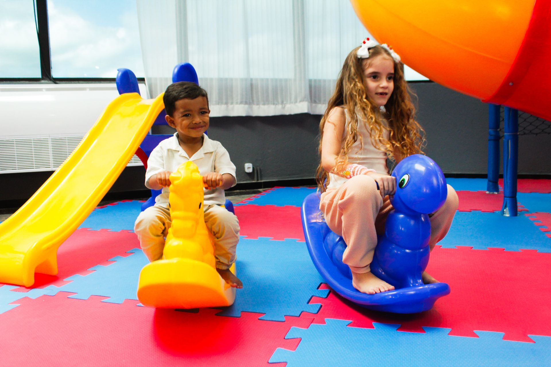 Duas crianças em brinquedos de balanço em uma área de recreação com escorregador e piso colorido.