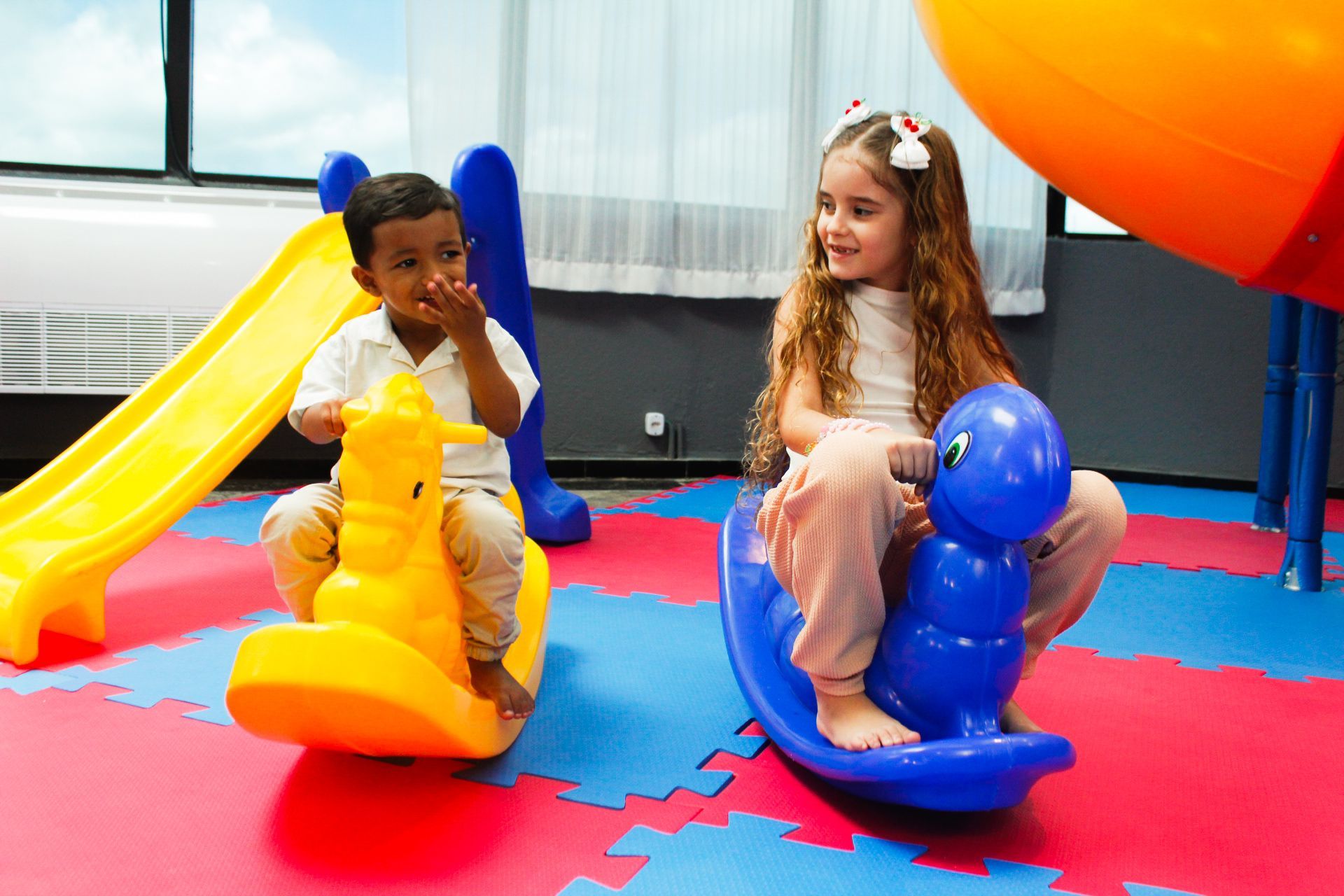 Duas crianças em brinquedos de balanço em uma área de recreação interna colorida.