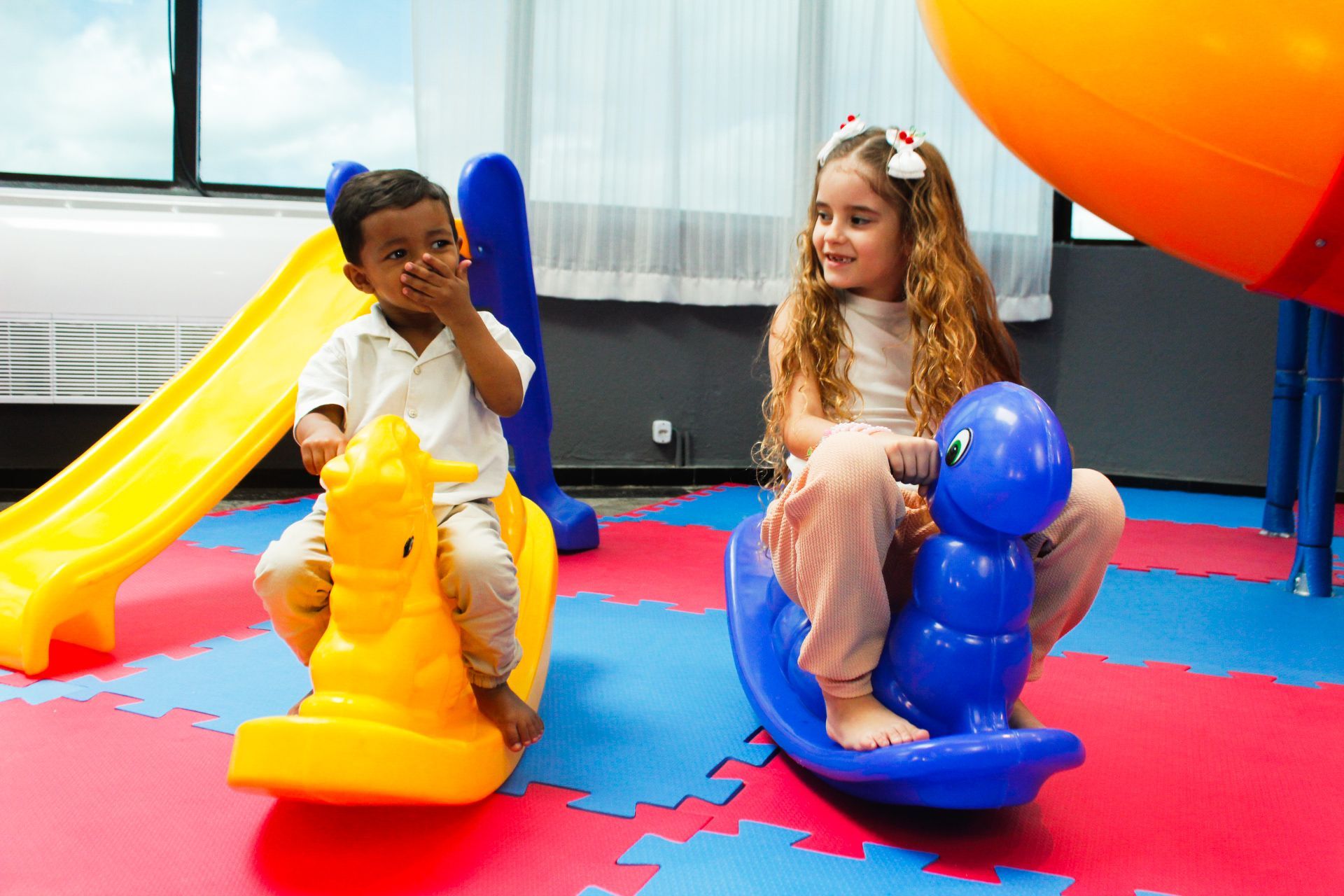 Menino e menina em brinquedos de balanço em uma área de recreação. Escorregador amarelo ao fundo.