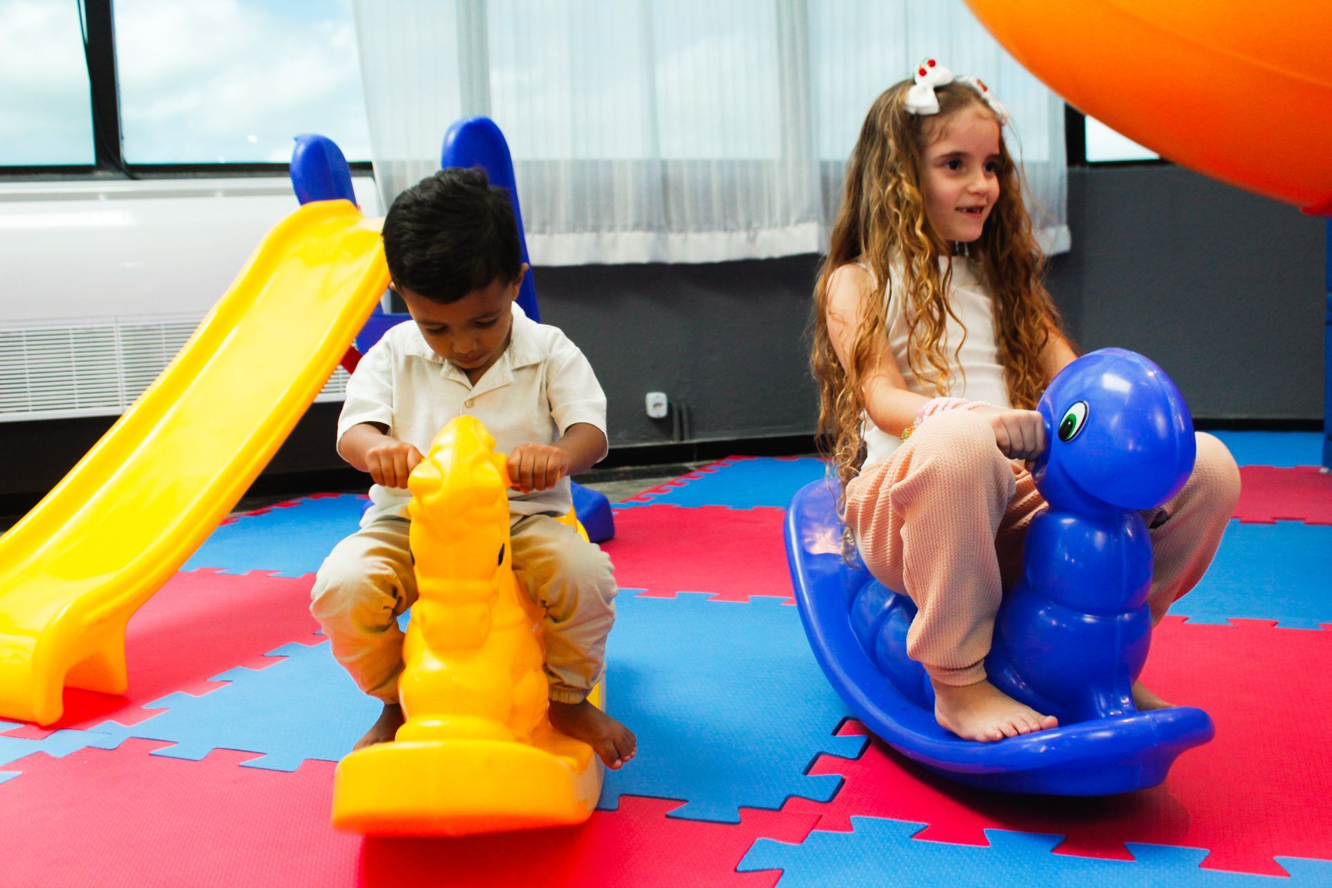 Duas crianças em brinquedos de balanço em uma área de recreação com escorregador; uma criança com uma camisa branca, a outra com um laço no cabelo.