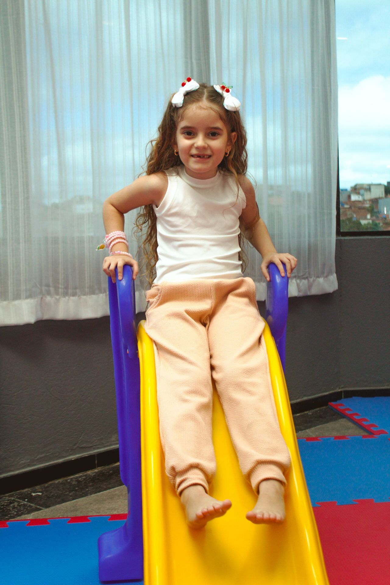 Menina deslizando por um escorregador amarelo e azul, sorrindo. Ambiente interno, perto da janela.