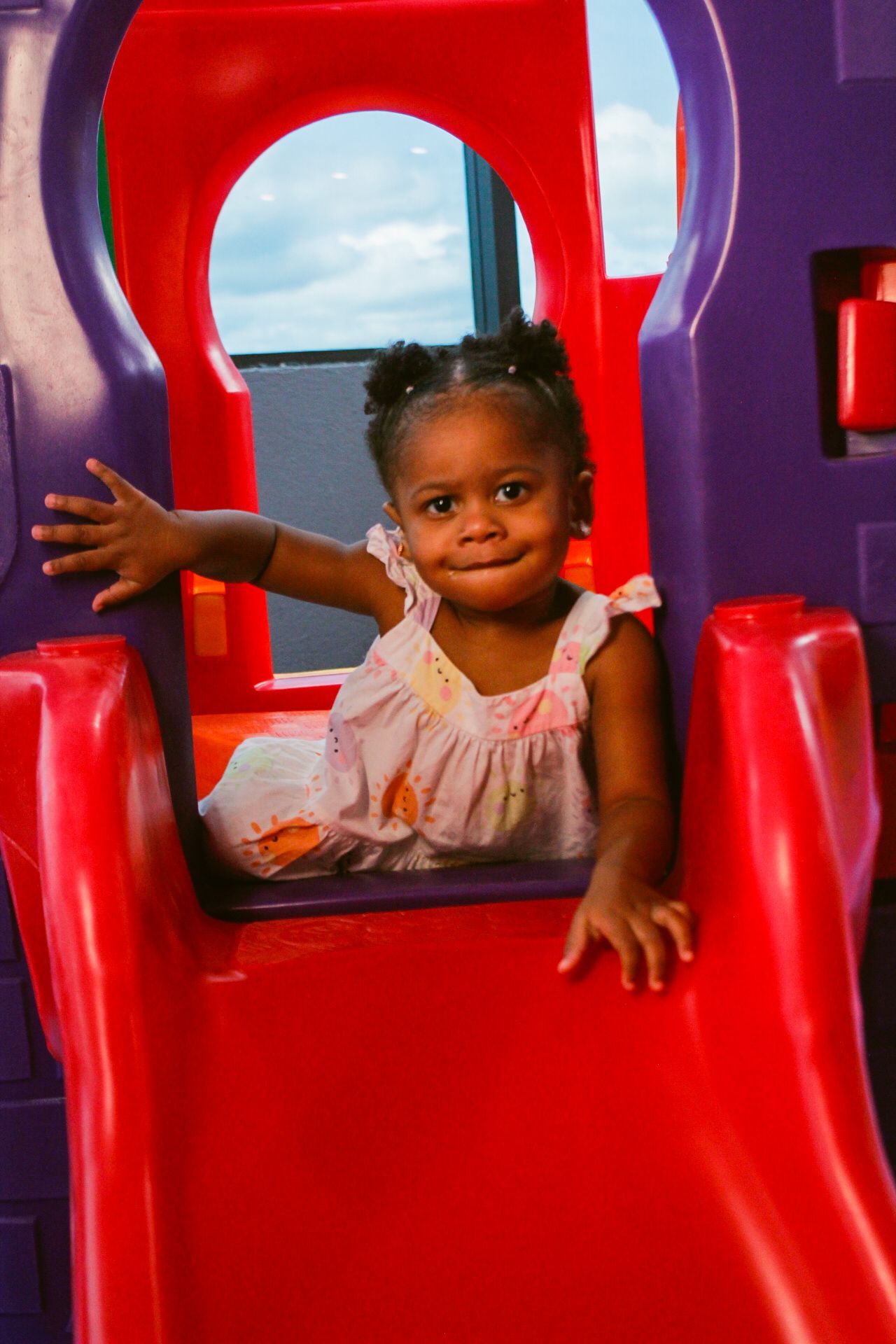 Criança em um escorregador vermelho e roxo, sorrindo e olhando para a câmera.
