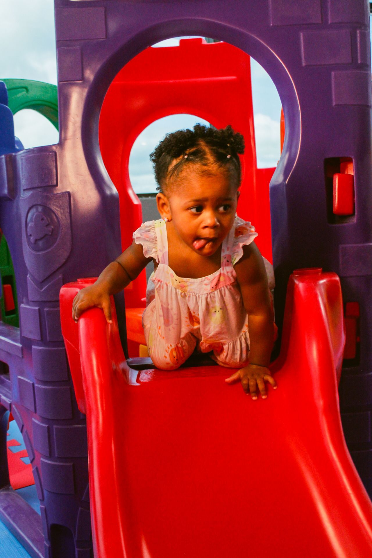 Criança em um escorregador vermelho, parte de um conjunto de brinquedos roxo e vermelho, mostrando a língua.