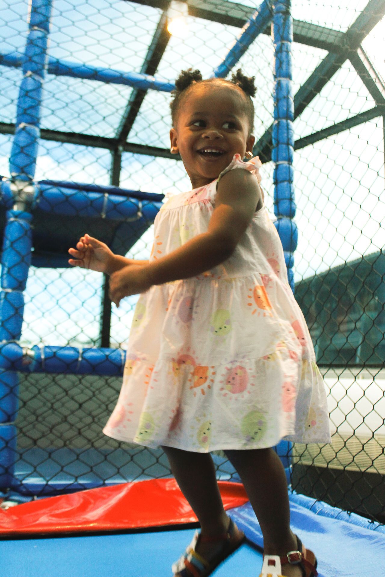 Criança sorridente de vestido branco pula em área de recreação interna com estruturas azuis e vermelhas.