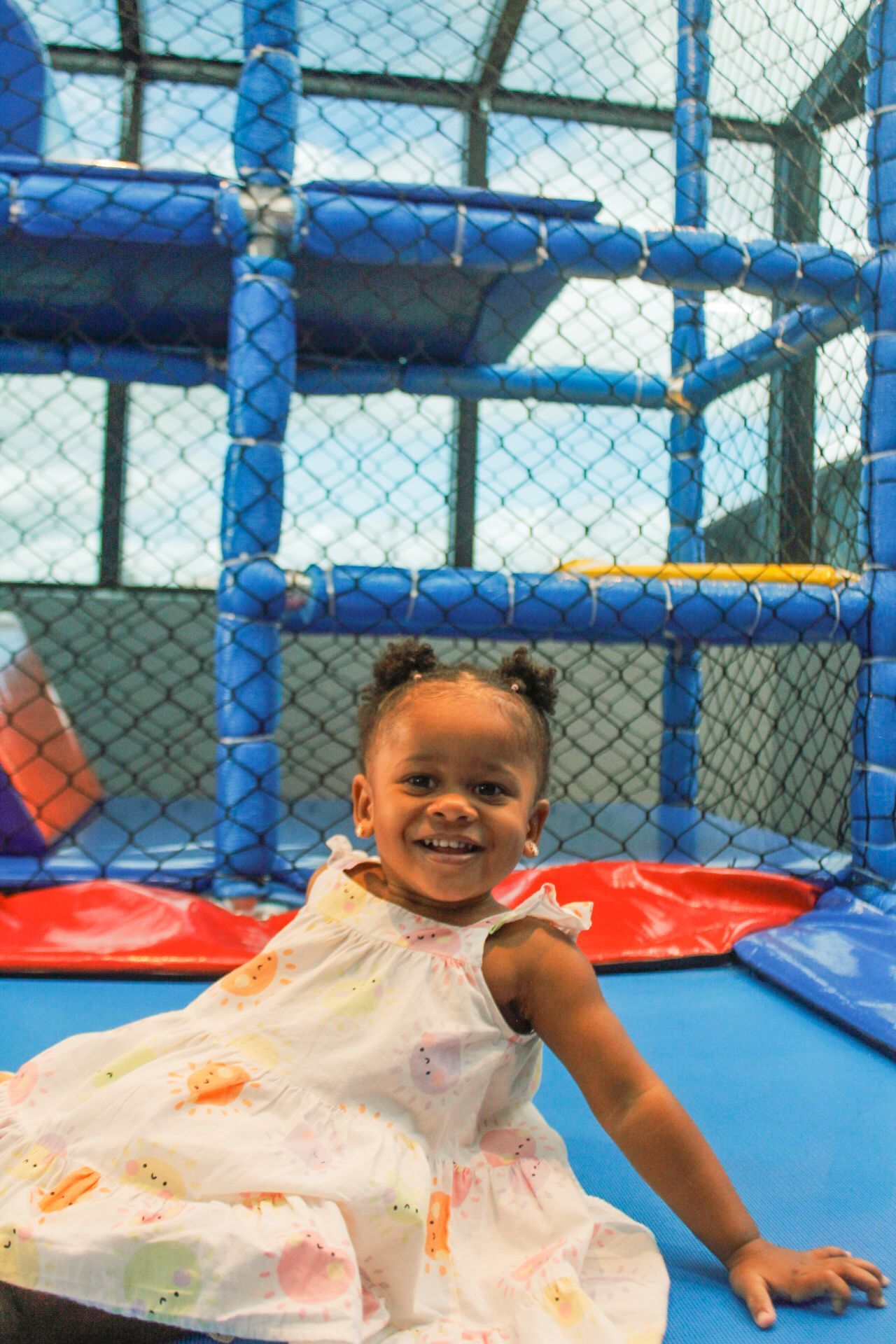 Criança pequena sorrindo, vestindo um vestido branco estampado, dentro de um parquinho infantil coberto em tons de azul e vermelho.
