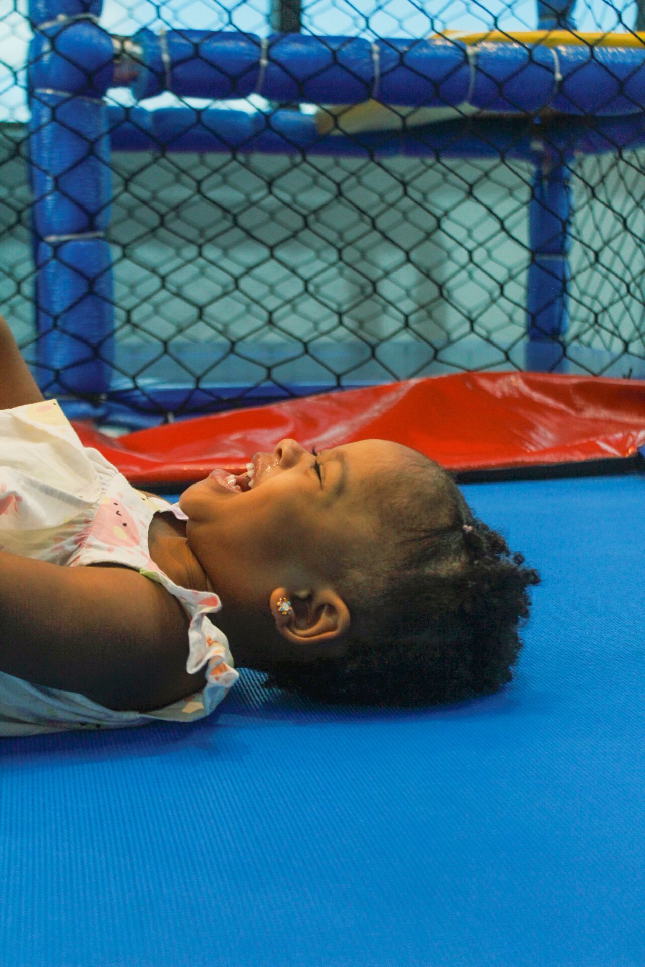 Criança rindo enquanto está deitada em uma superfície acolchoada azul, plataforma vermelha, com estrutura de brinquedo azul e preta ao fundo.