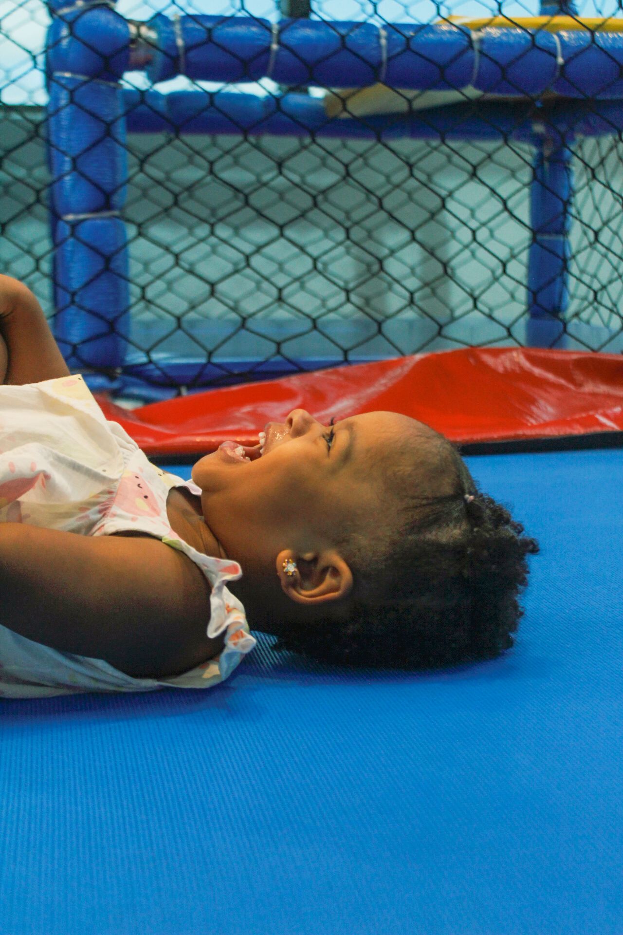 Criança rindo, deitada em um tapete azul, com uma área de recreação cercada por rede ao fundo.