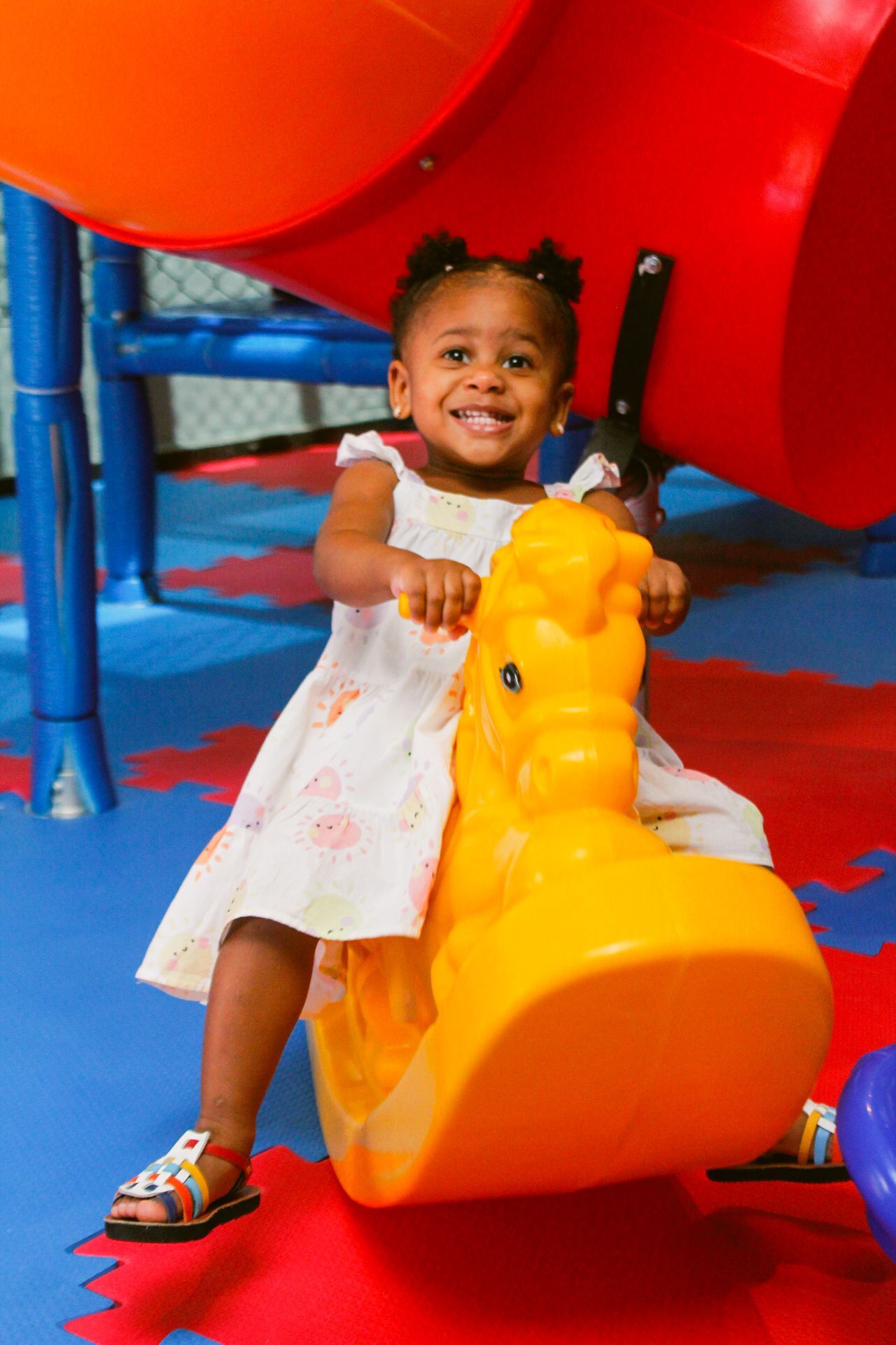 Menina sorridente em um cavalinho de balanço amarelo, em uma área de recreação colorida com um escorregador.
