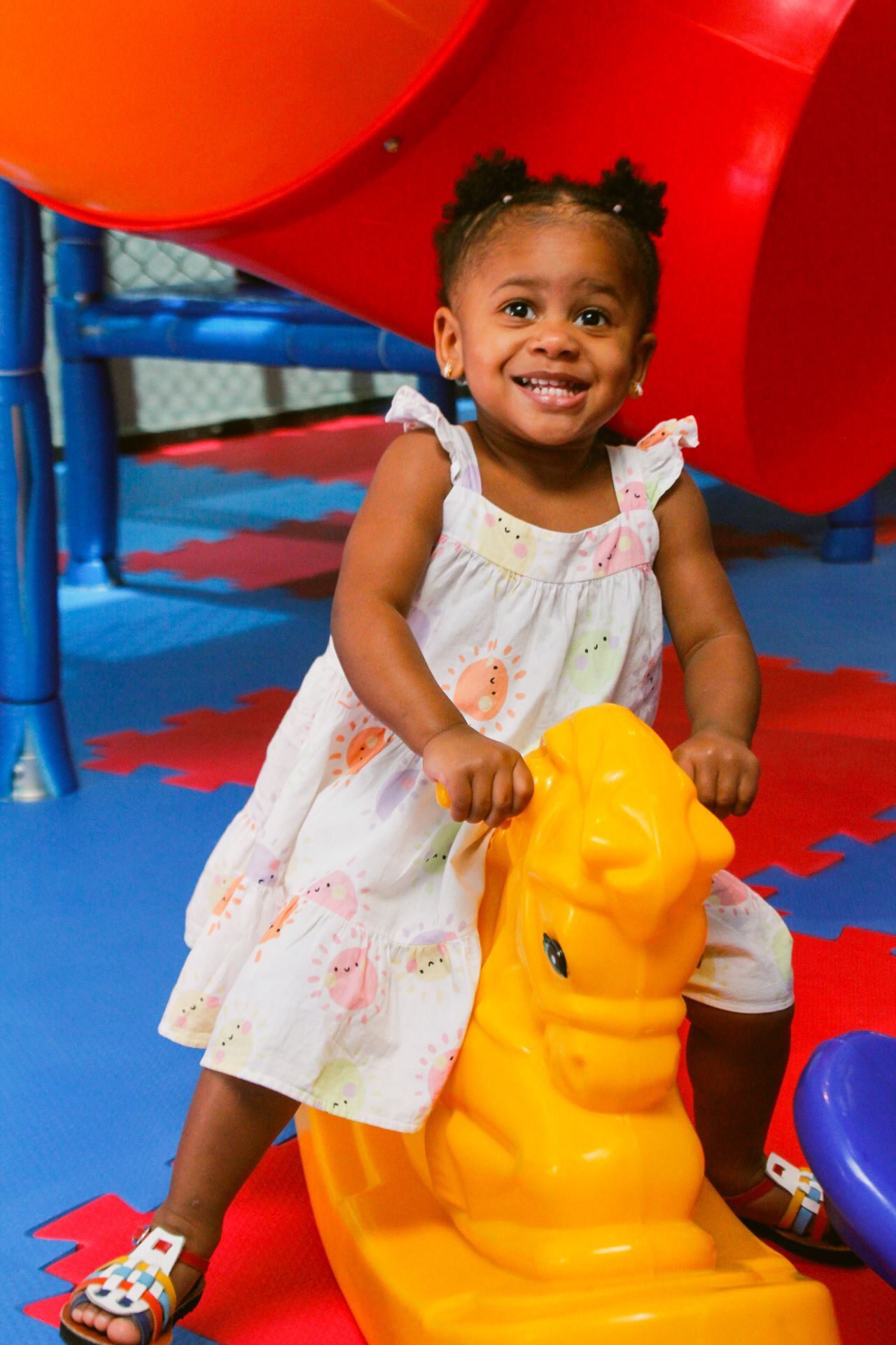 Criança sorrindo enquanto monta um cavalinho de balanço amarelo, em uma área de recreação com escorregadores coloridos.