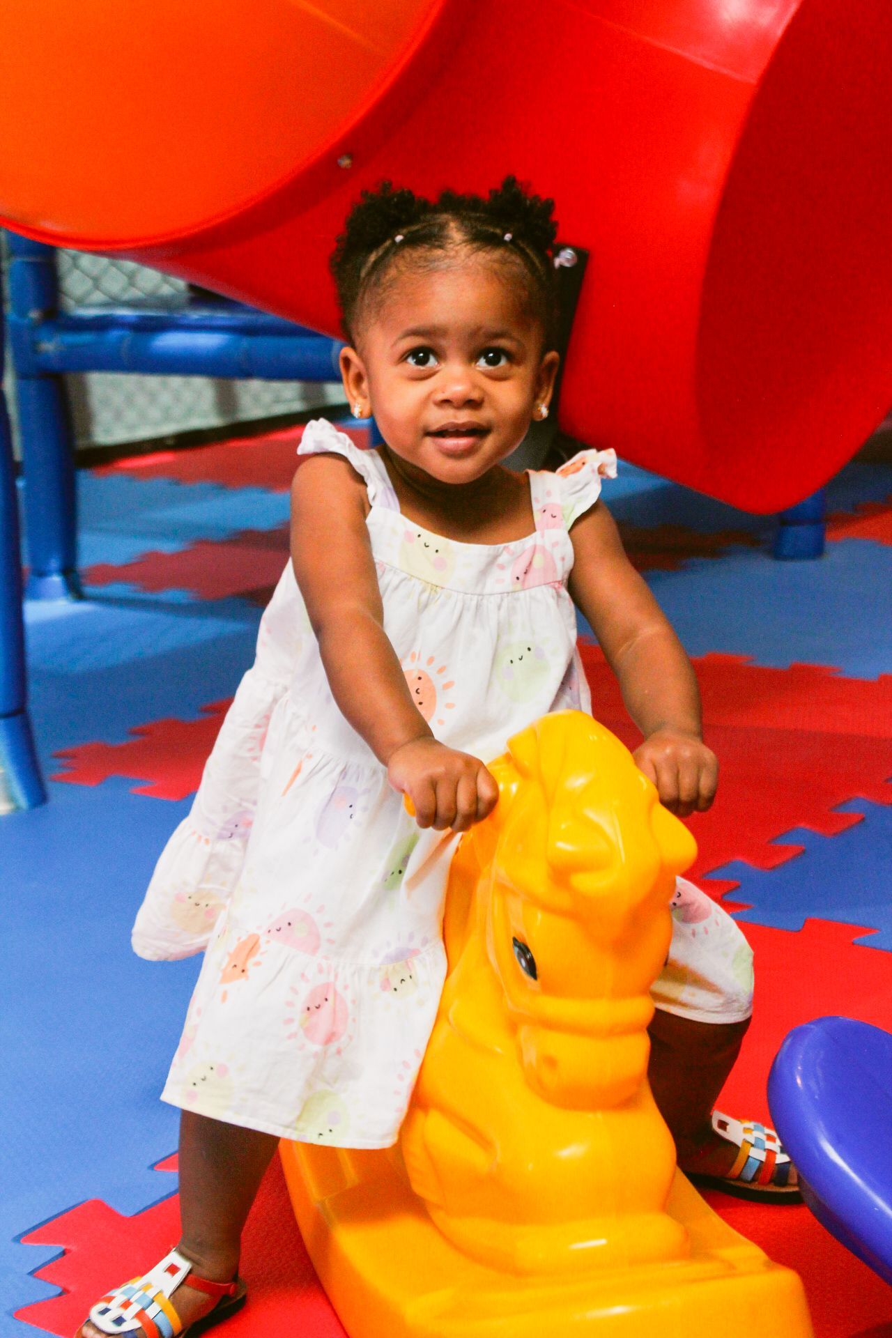 Criança pequena sorrindo em um cavalinho de balanço amarelo, dentro de um colorido parque infantil coberto.