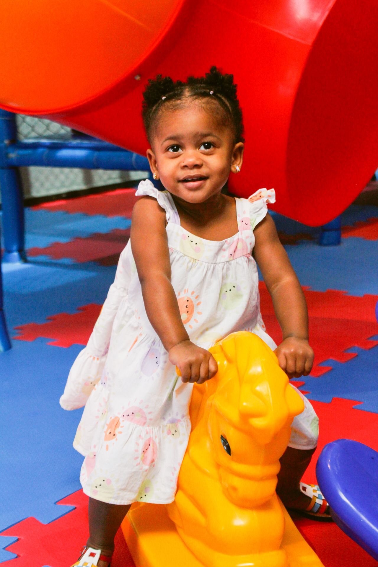Criança pequena sorrindo em um cavalinho de brinquedo amarelo, em uma área de recreação colorida.