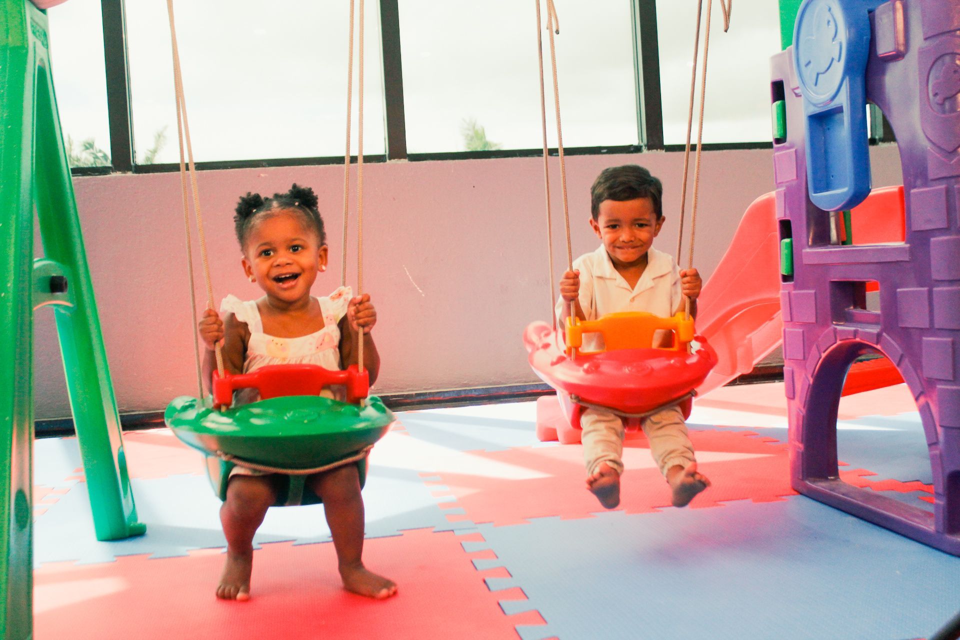 Duas crianças sorrindo em balanços dentro de casa, perto de um parquinho infantil.
