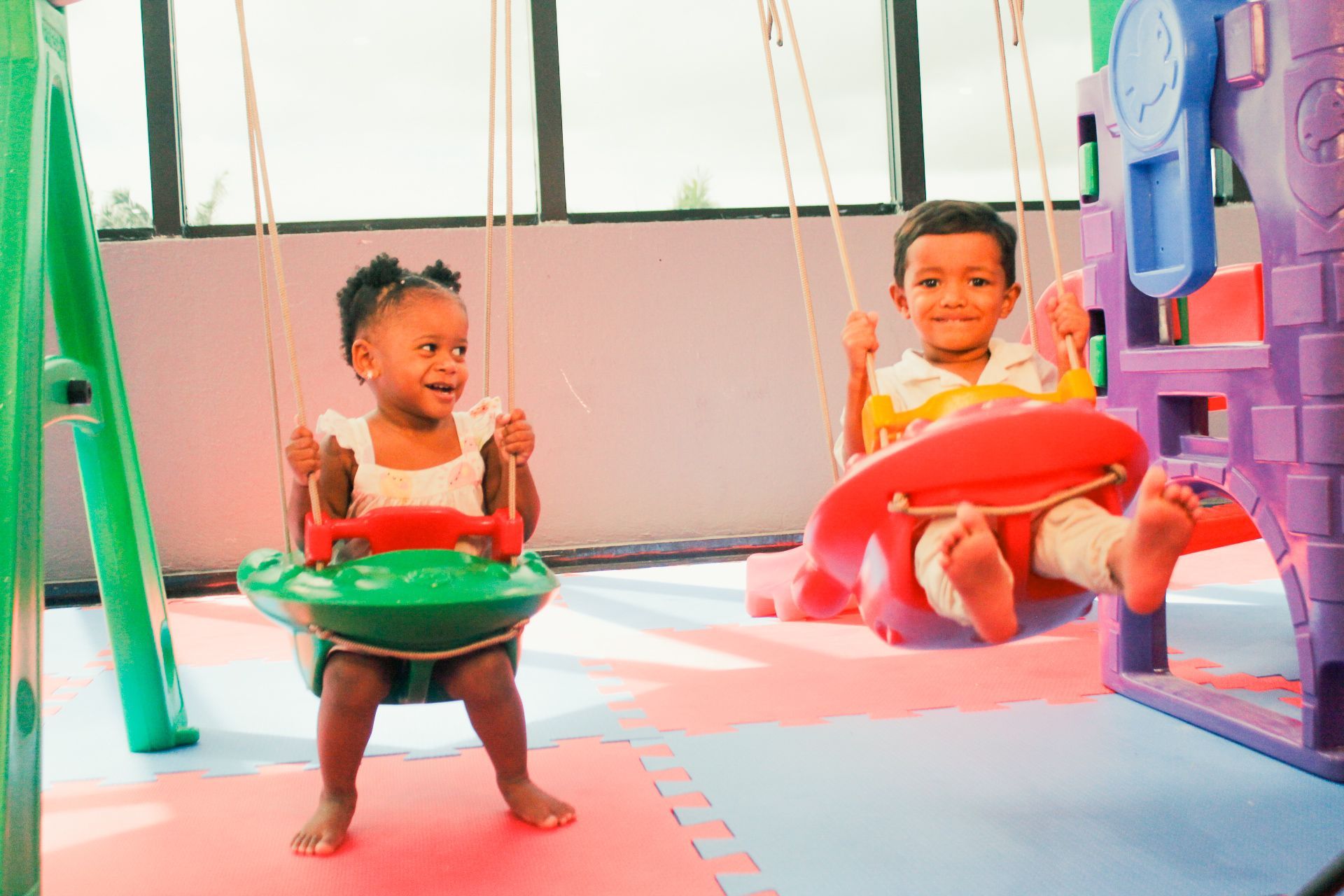 Duas crianças sorridentes em balanços numa área de recreação. Balanços vermelhos e verdes, estrutura de brinquedos colorida.