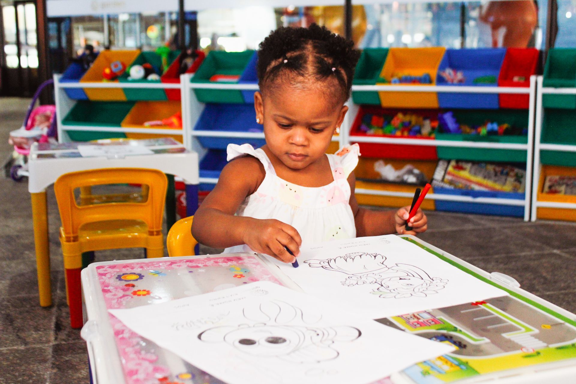 Criança pequena colorindo em uma mesa com brinquedos e prateleiras ao fundo.