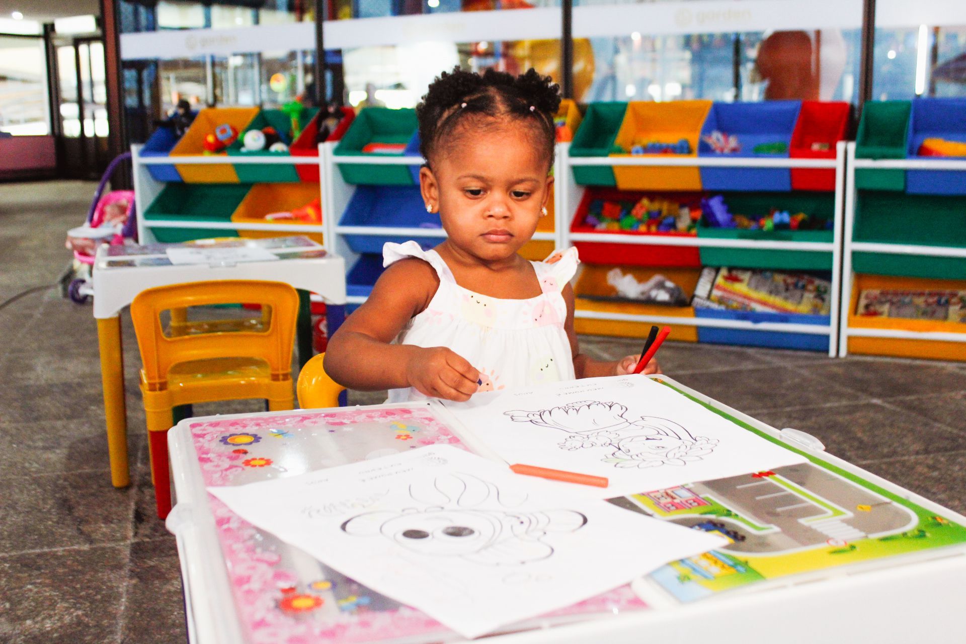 Menina colorindo em uma mesa com brinquedos e prateleiras em uma sala de jogos colorida.