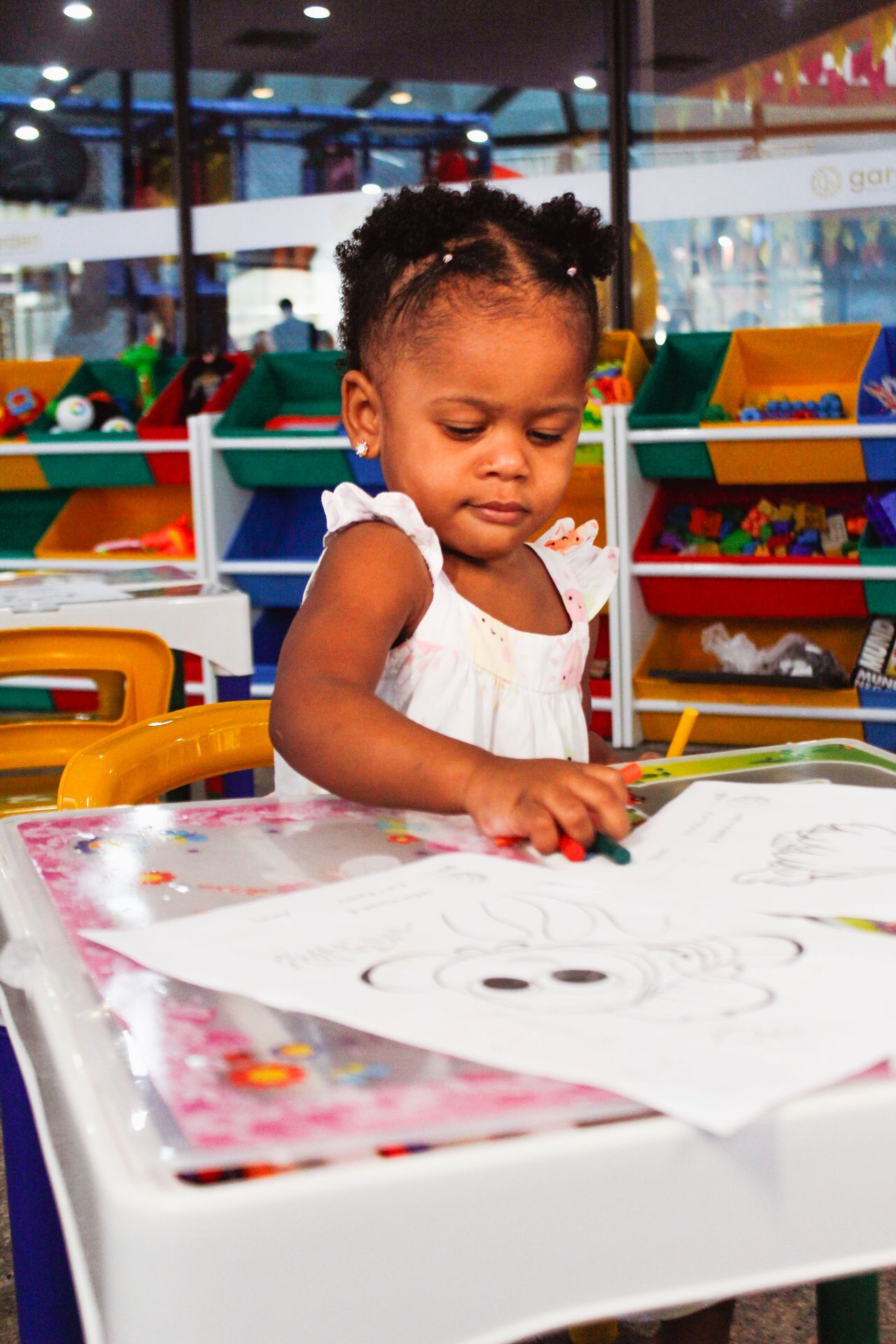 Criança pequena colorindo em uma mesa na sala de jogos, concentrada no desenho com giz de cera.
