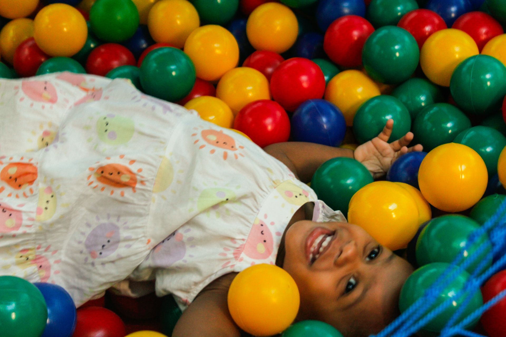 Criança sorrindo, deitada em uma piscina de bolinhas colorida com bolinhas vermelhas, amarelas, verdes e azuis.