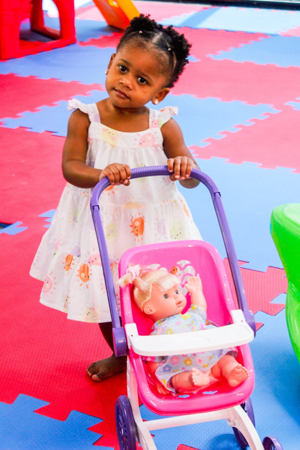 Menina empurrando um carrinho de boneca rosa, com uma boneca dentro, sobre um tapete de brincar colorido.