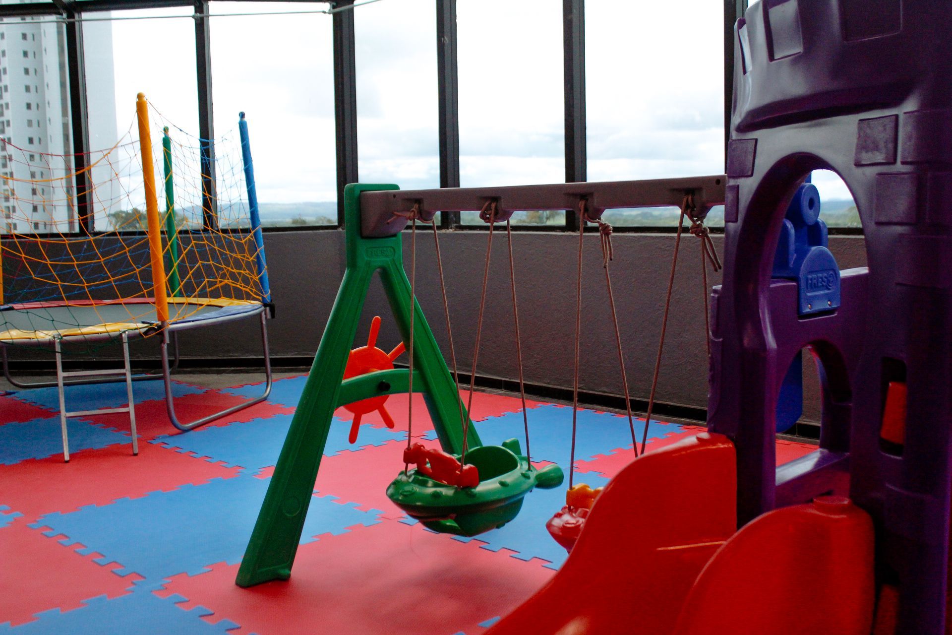 Parque infantil coberto com balanço, cama elástica e escorregador, sobre tapetes azuis e vermelhos.