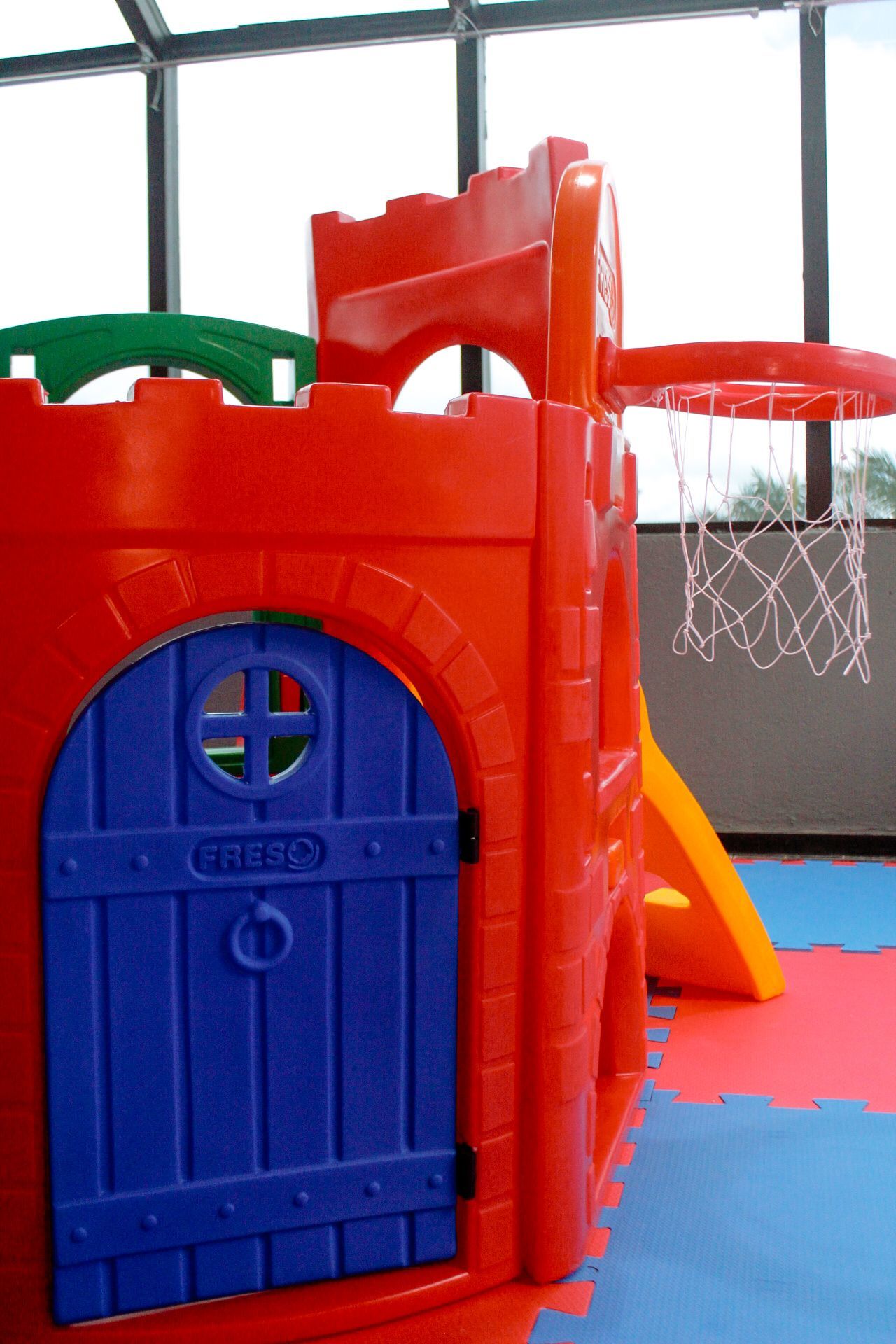 Castelo de plástico vermelho e azul com cesta de basquete, dentro de uma sala bem iluminada.